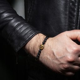 Photo & Brand - Fotografia, Strony WWW - Męska bransoletka z czarnych kamieni lawowych i złotą ozdobą czaszki na nadgarstku w skórzanej kurtce, w ciemnym otoczeniu. Detal produktu.