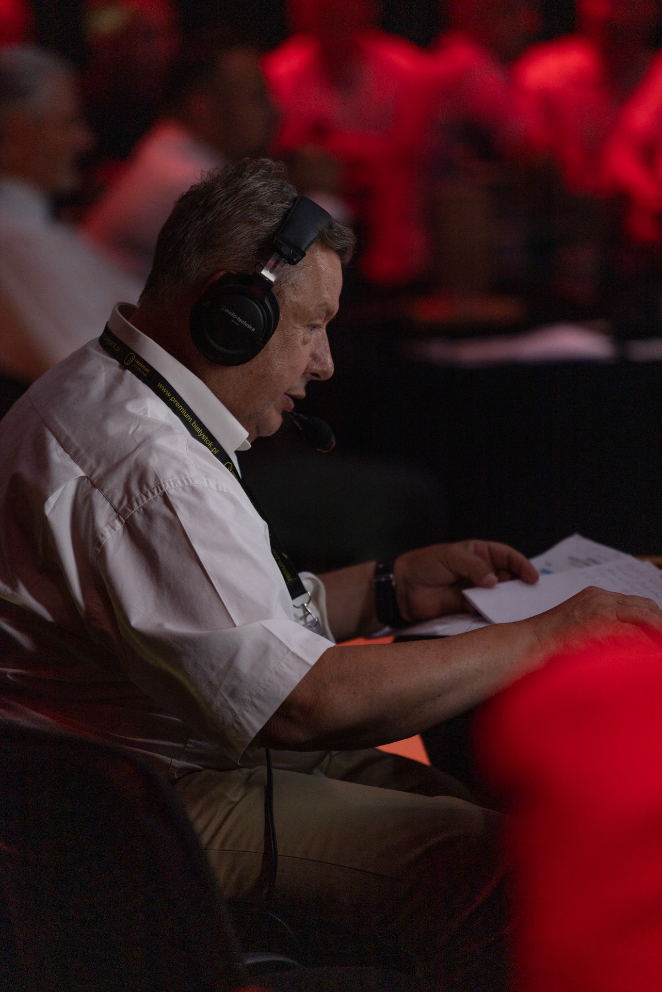 Mężczyzna w słuchawkach z mikrofonem, w białej koszuli, przegląda dokumenty podczas wydarzenia oświetlonego czerwonym światłem.