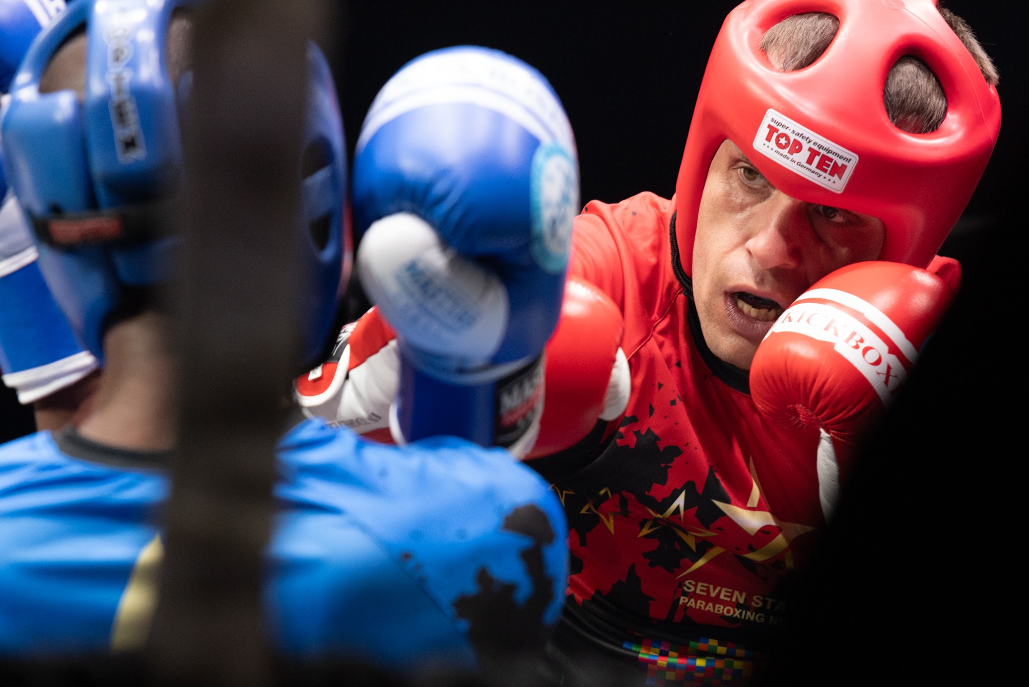 Zawodnik kickboxingu w czerwonym ochraniaczu na głowę i czerwonych rękawicach, w trakcie walki z przeciwnikiem w niebieskim ochraniaczu i niebieskich rękawicach.