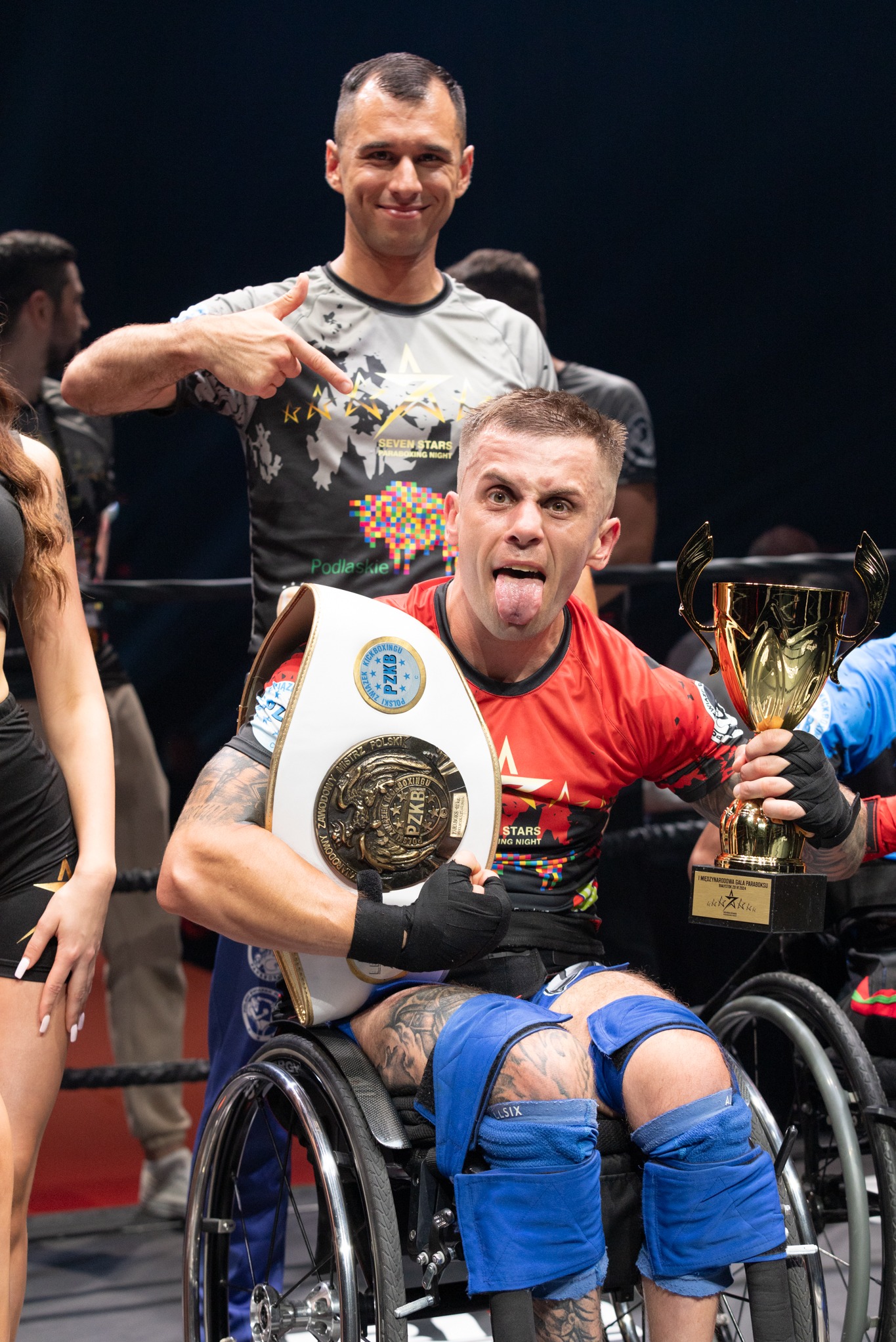 Uśmiechnięty sportowiec na wózku inwalidzkim, z wyciągniętym językiem, trzyma złoty puchar i pas mistrzowski PZKB, w tle mężczyzna wskazuje palcem na logo 'Seven Stars Paraboxing Night'.