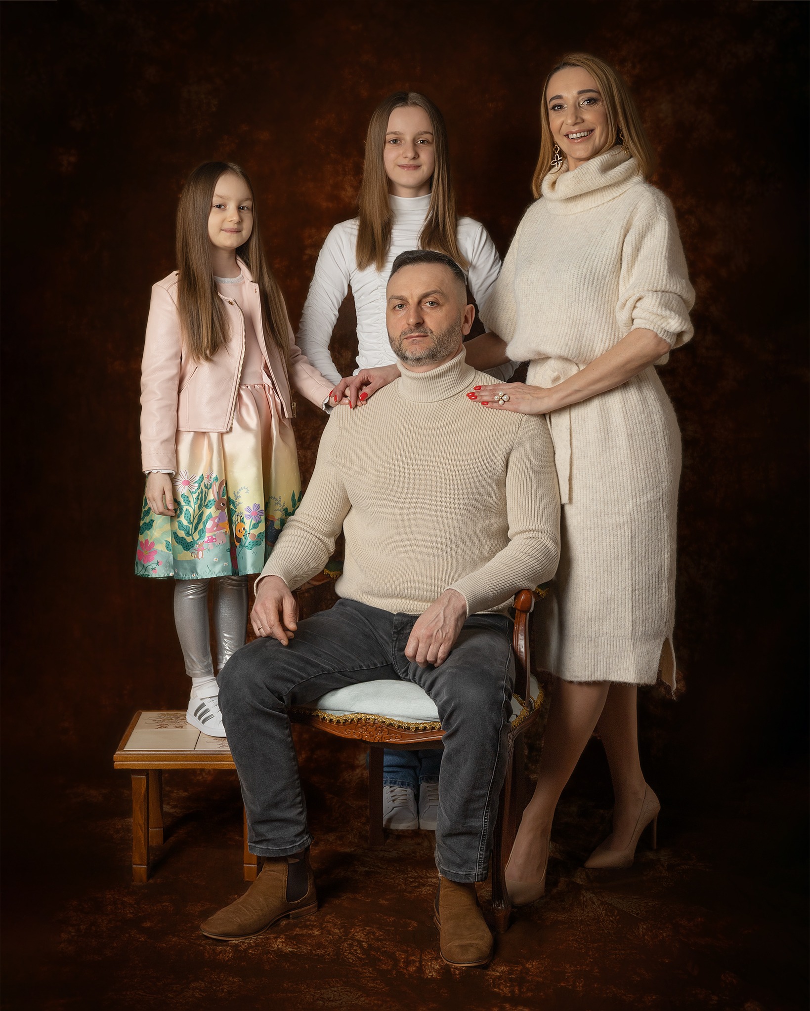 Portret rodzinny: ojciec siedzi na krześle, matka i córki stoją za nim, na brązowym tle. Matka z ręką na ramieniu ojca, młodsza córka w różowej kurtce i sukience z motywem leśnym, starsza w białym...