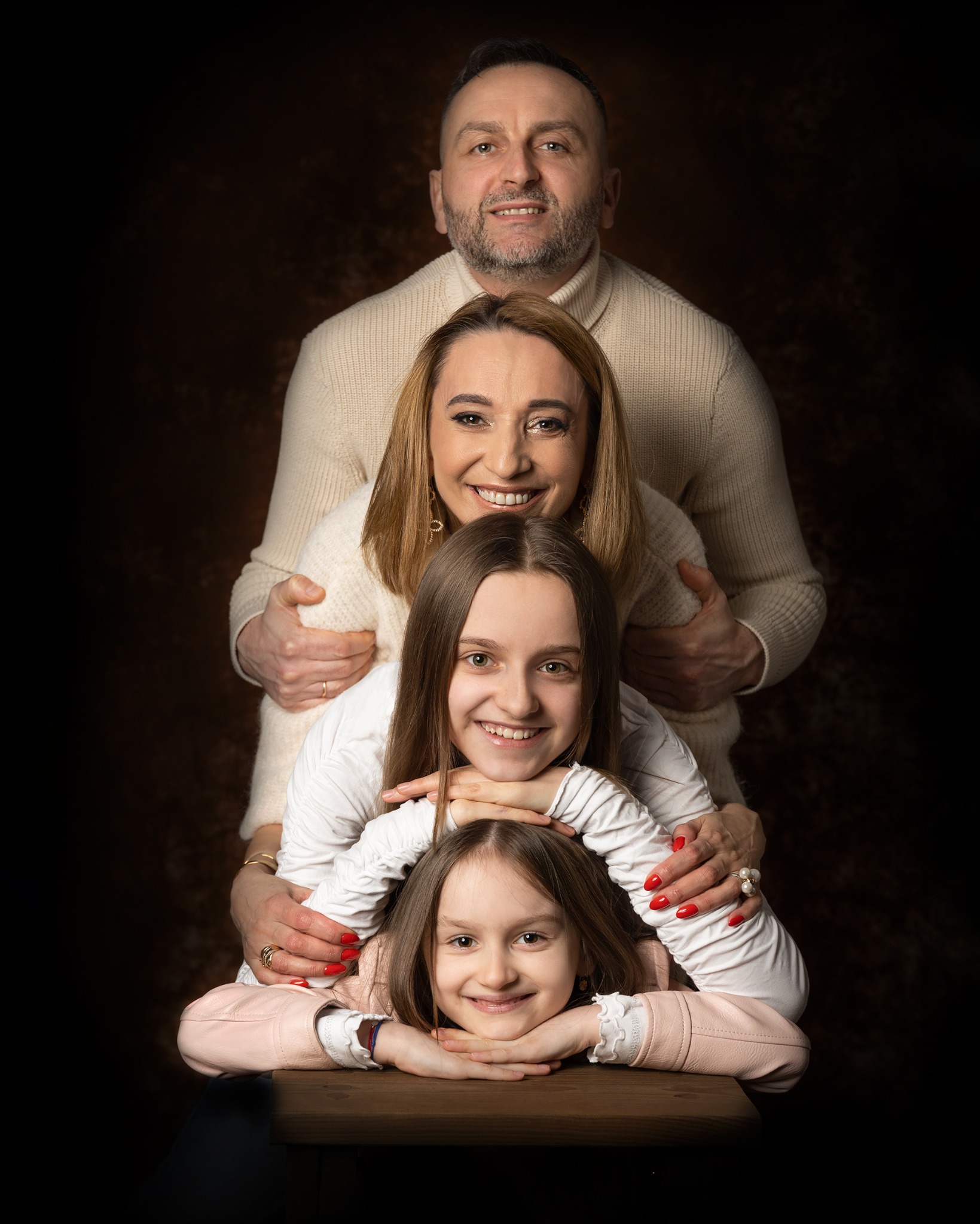 Portret rodzinny w studio: ojciec obejmujący matkę, dwie córki ułożone jedna pod drugą, uśmiechnięte twarze, ciemne tło z subtelną teksturą.