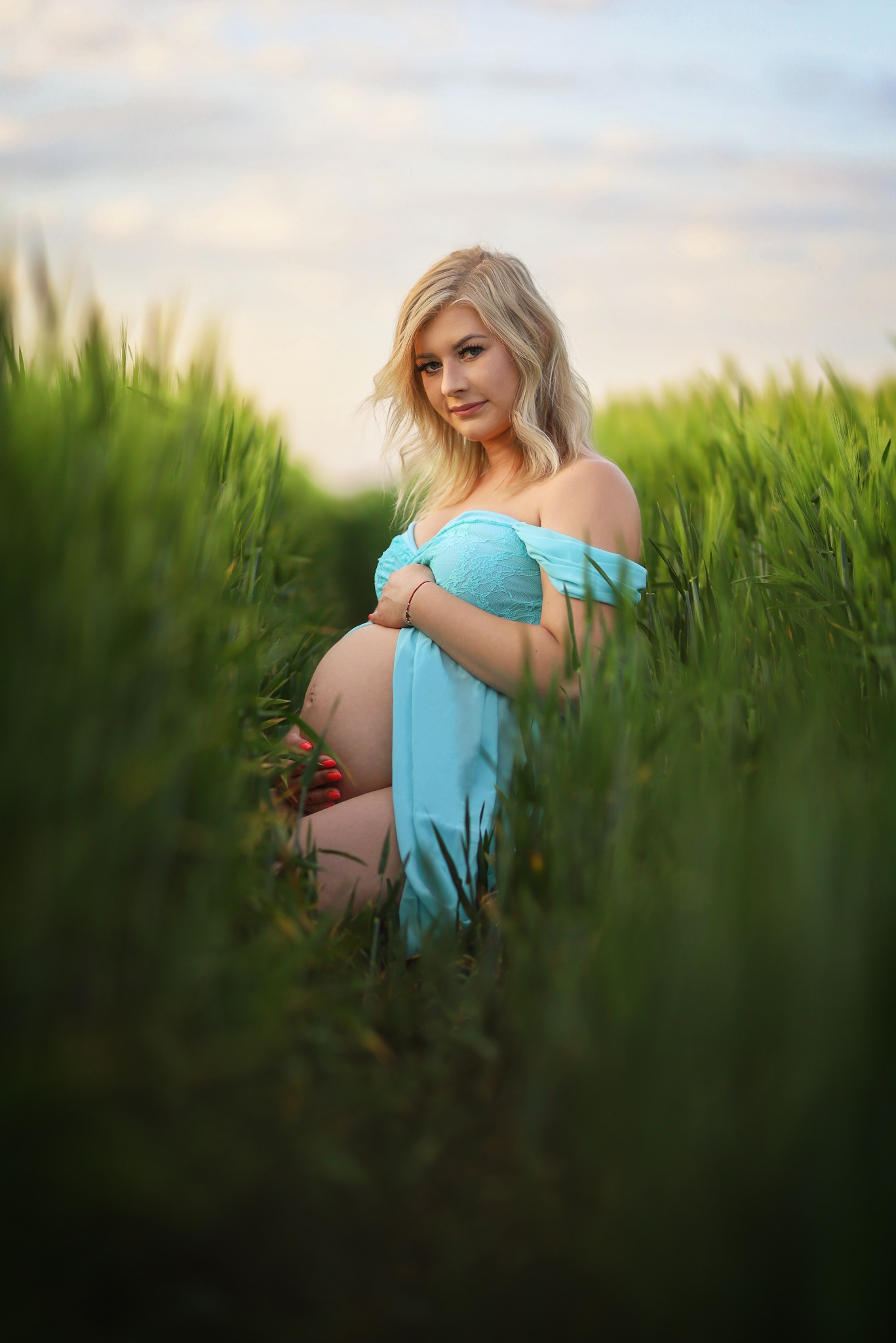 Kobieta w ciąży, ubrana w błękitną sukienkę, pozuje siedząc w wysokiej trawie, z dłońmi ułożonymi na brzuchu i delikatnym uśmiechem.
