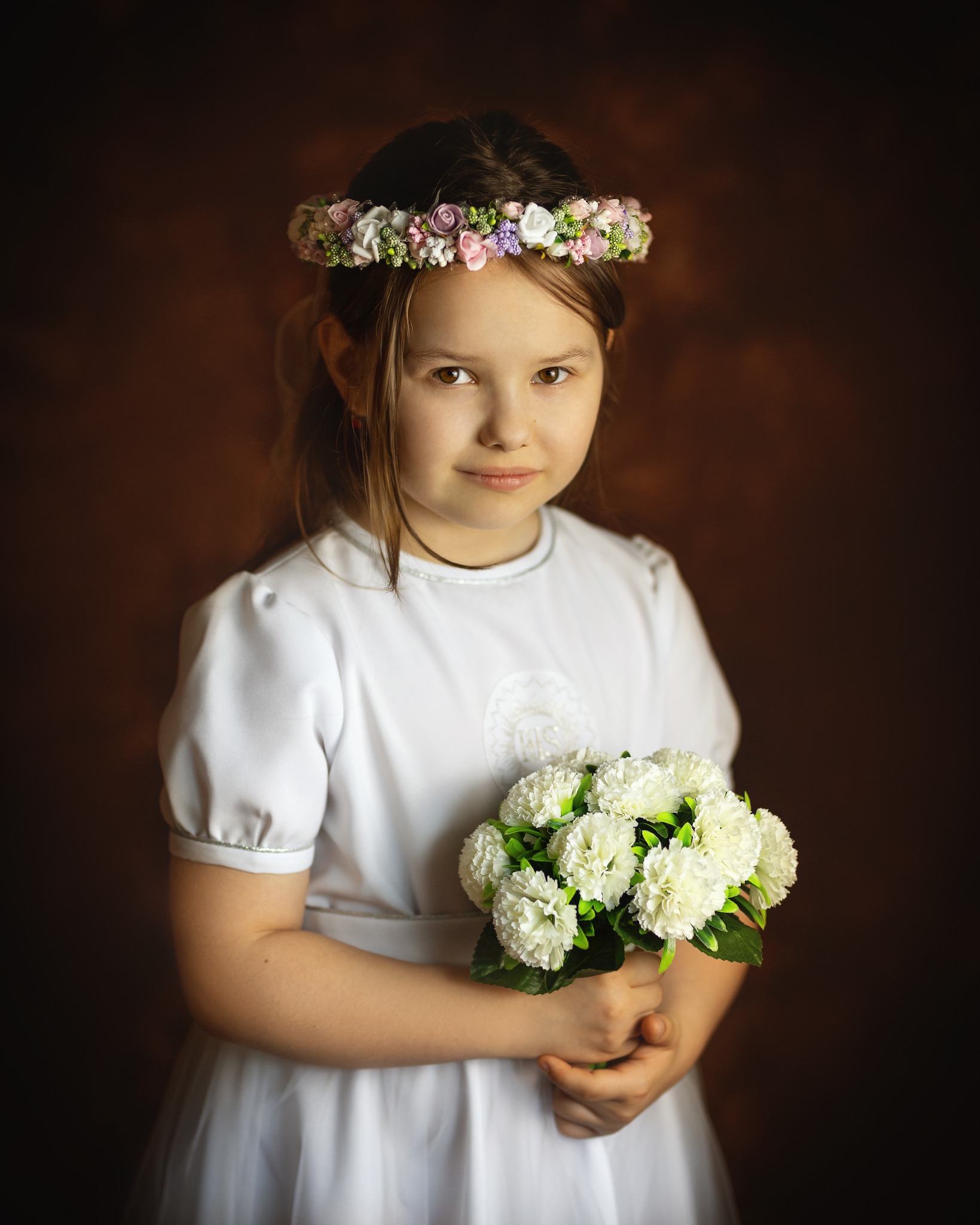 Portret dziewczynki w białej sukience komunijnej z wiankiem z kwiatów na głowie, trzymającej bukiet białych kwiatów na ciemnobrązowym tle.