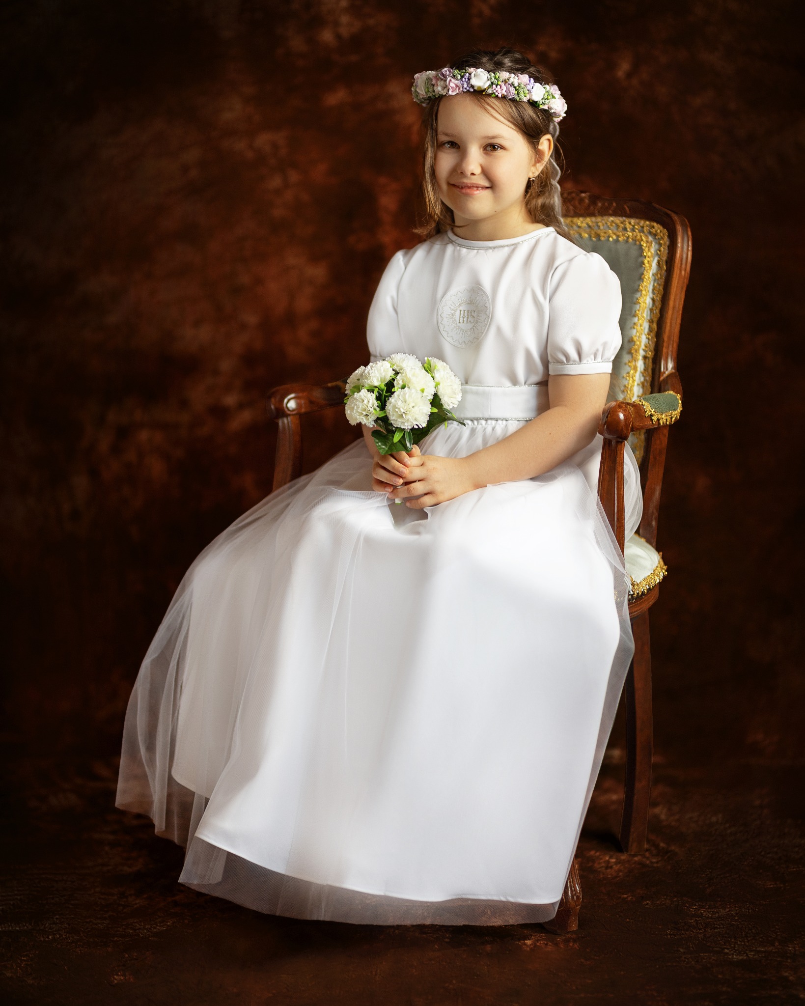 Dziewczynka w białej sukience komunijnej z wiankiem na głowie, siedząca na zdobionym krześle i trzymająca bukiecik białych kwiatów na tle brązowego tła.