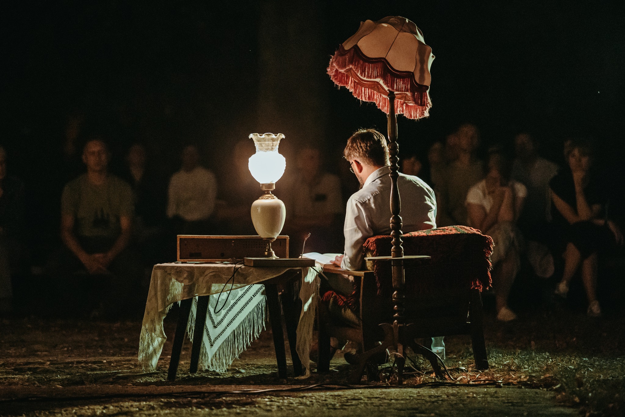 Mężczyzna w koszuli siedzi przy stole oświetlonym lampą, pisząc coś na kartce podczas występu na scenie przed publicznością w plenerze.