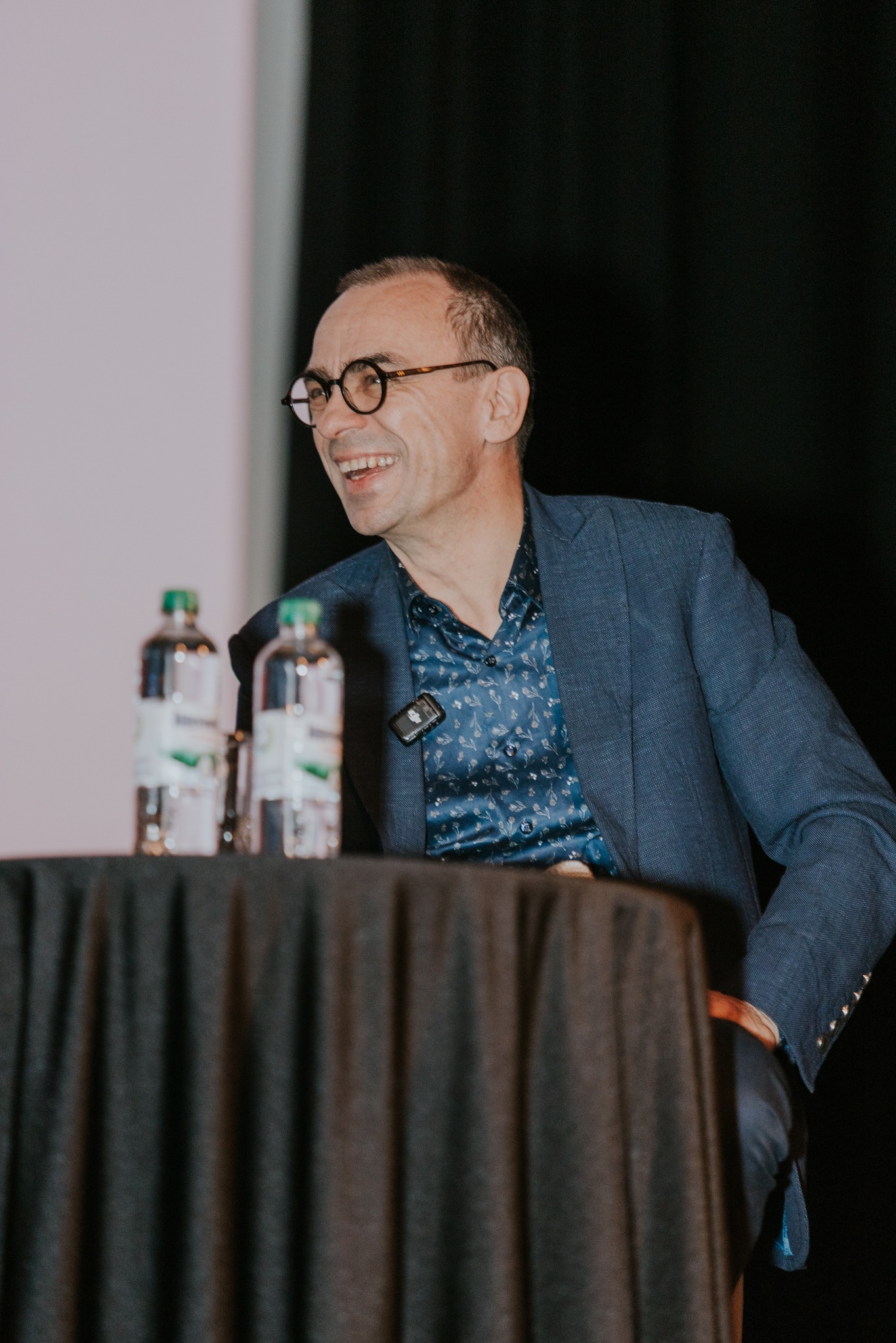 Mężczyzna w okularach i niebieskiej marynarce uśmiecha się podczas przemówienia na tle sceny z dwoma butelkami wody na stole.