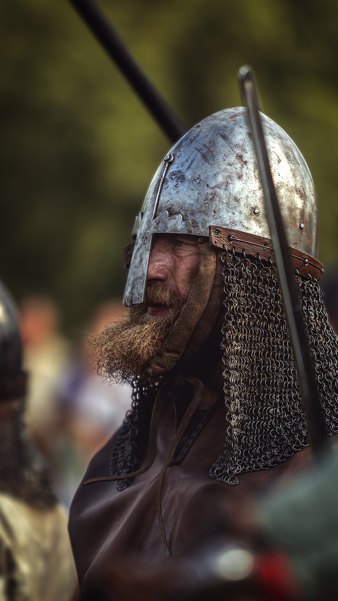 Portret brodatego mężczyzny w hełmie i kolczudze, z profilu. Widoczne detale stroju wojownika, zarys twarzy i faktura metalu. Tło rozmyte, plenerowe.