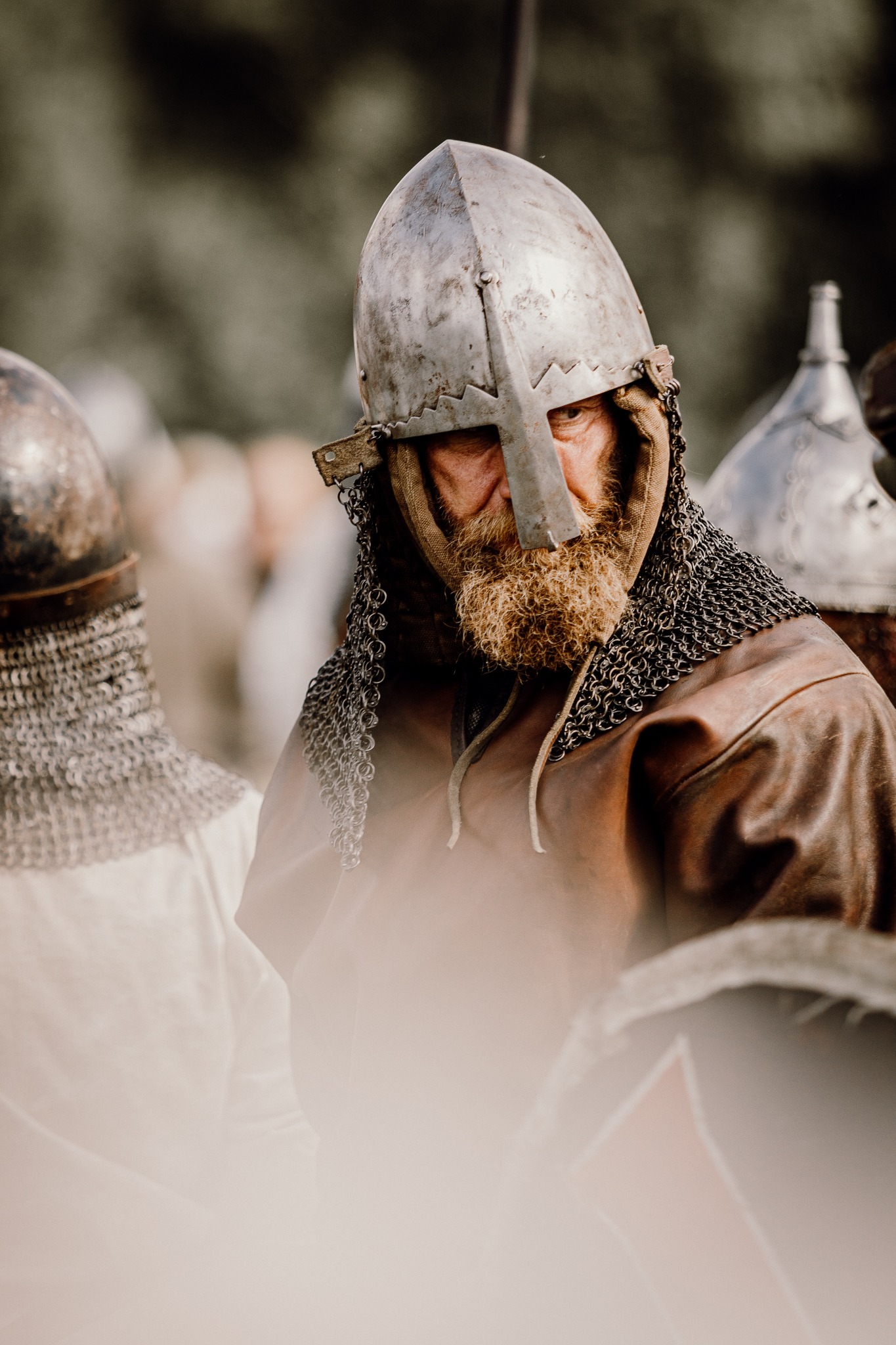 Portret brodatego mężczyzny w hełmie wikinga z kolczugą, na tle innych wojowników. Ujęcie z bliska, skupione na twarzy i detalu stroju.