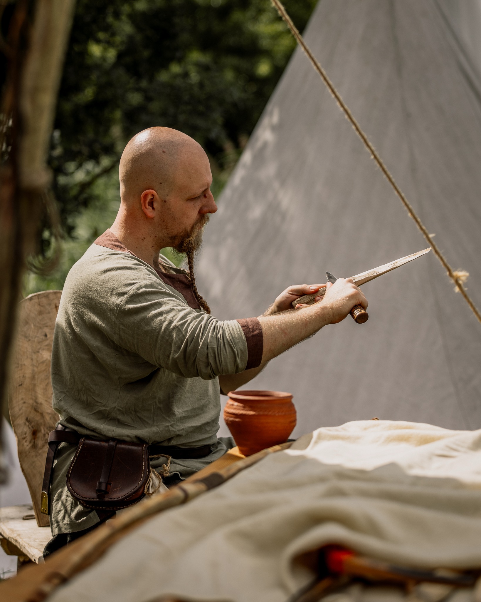 Mężczyzna w stroju historycznym rzeźbi w drewnie nożem. Widoczny gliniany garnek i skórzana torba. W tle namiot i zieleń.