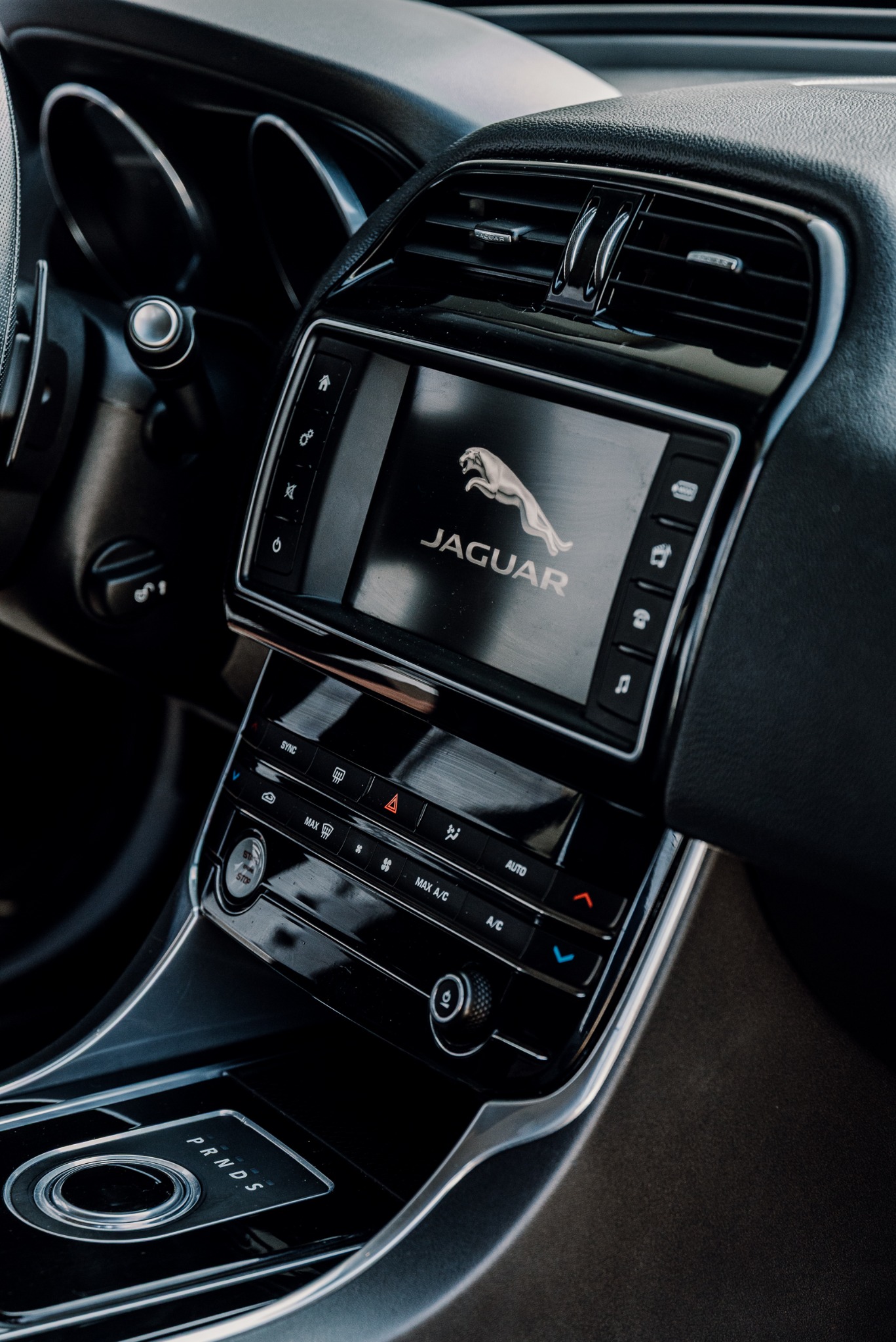 Wnętrze luksusowego samochodu Jaguar, z widocznym panelem sterowania, ekranem z logo marki i elementami deski rozdzielczej. Dominują ciemne kolory i elegancki design.