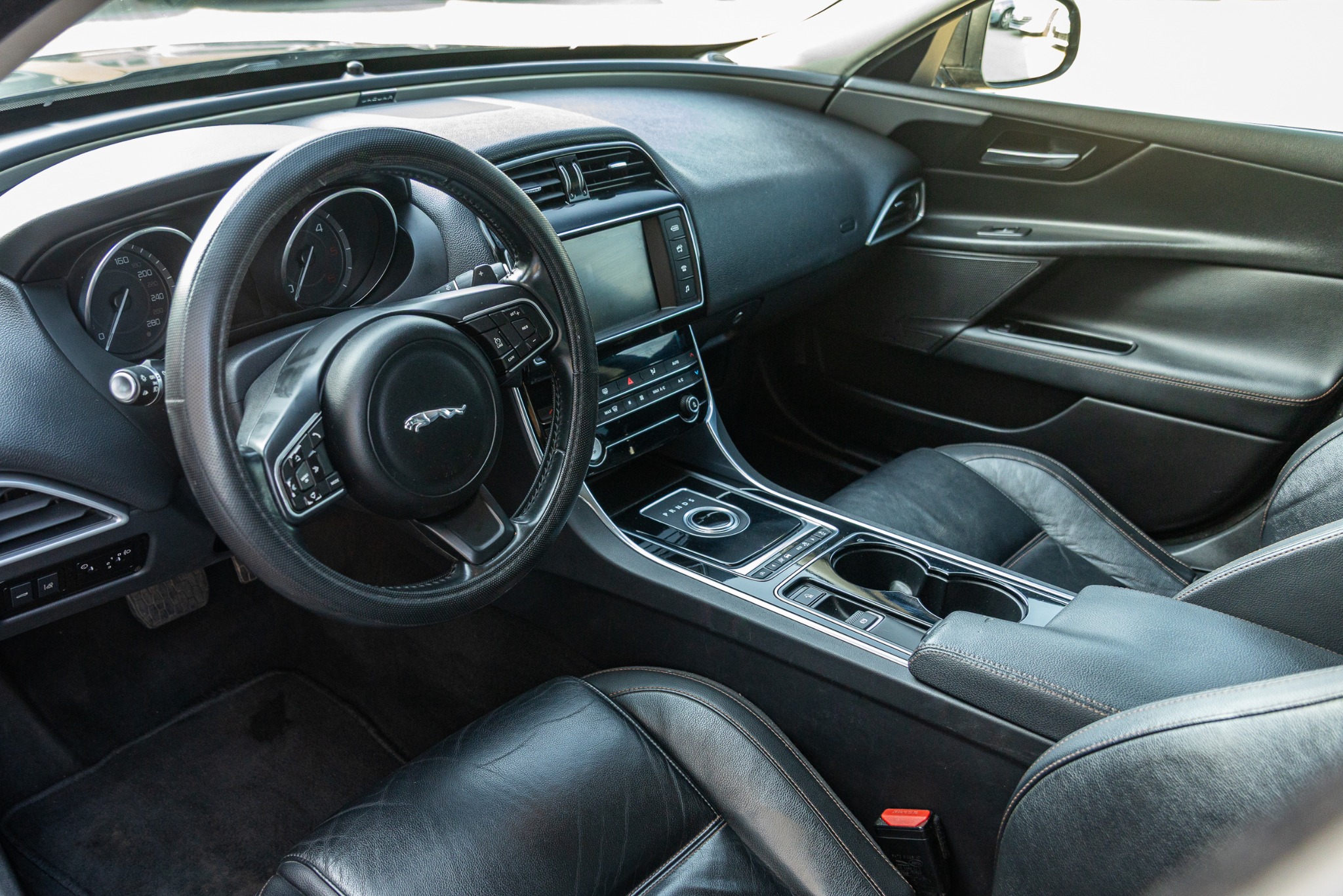 Wnętrze luksusowego samochodu Jaguar z czarną skórzaną tapicerką, deską rozdzielczą i kierownicą. Widoczne detale wykończenia i elementy sterowania.