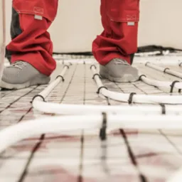 Instalacja ogrzewania podłogowego: widok z dołu na białe rurki mocowane czarnymi uchwytami do siatki, pracownik w czerwonych spodniach roboczych i szarych butach oraz narzędzie do montażu.
