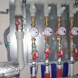 Rozdzielacz instalacji grzewczej z czterema licznikami ciepła, zaworami i izolacją termiczną rur, zamontowany na metalowej płycie.