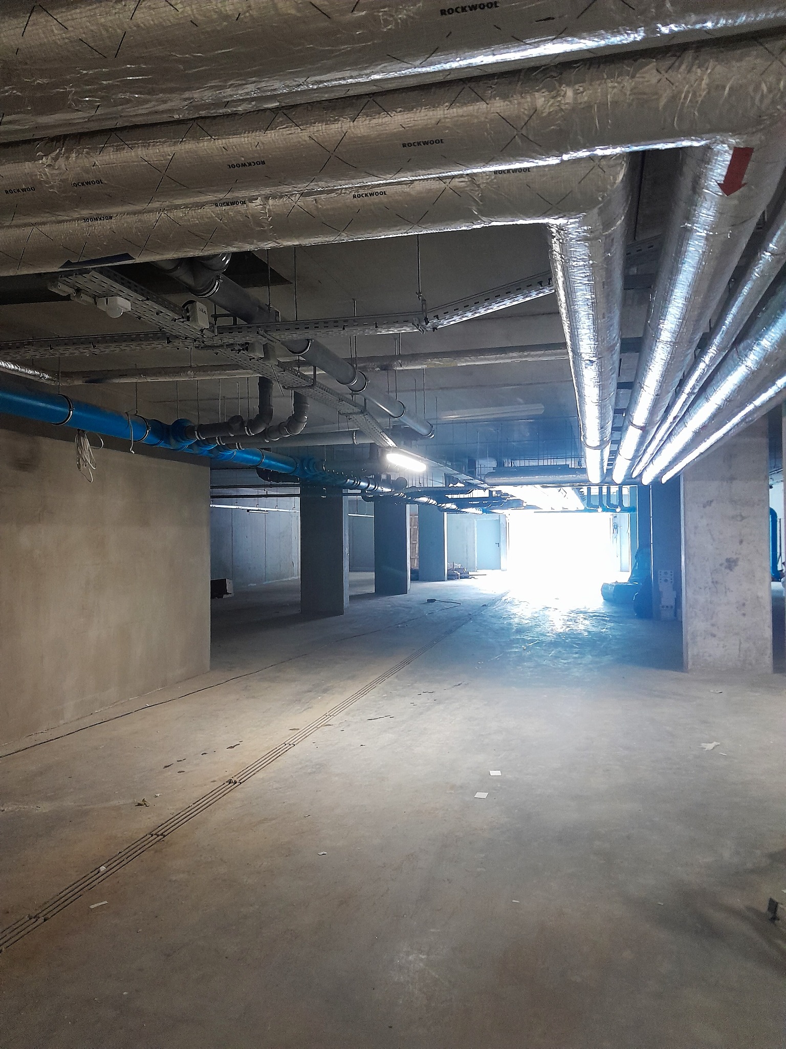 Instalacja rur wentylacyjnych i hydraulicznych pod sufitem w surowym wnętrzu, z widocznymi izolacjami Rockwool i niebieskimi rurami, oświetlona odległym światłem dziennym.