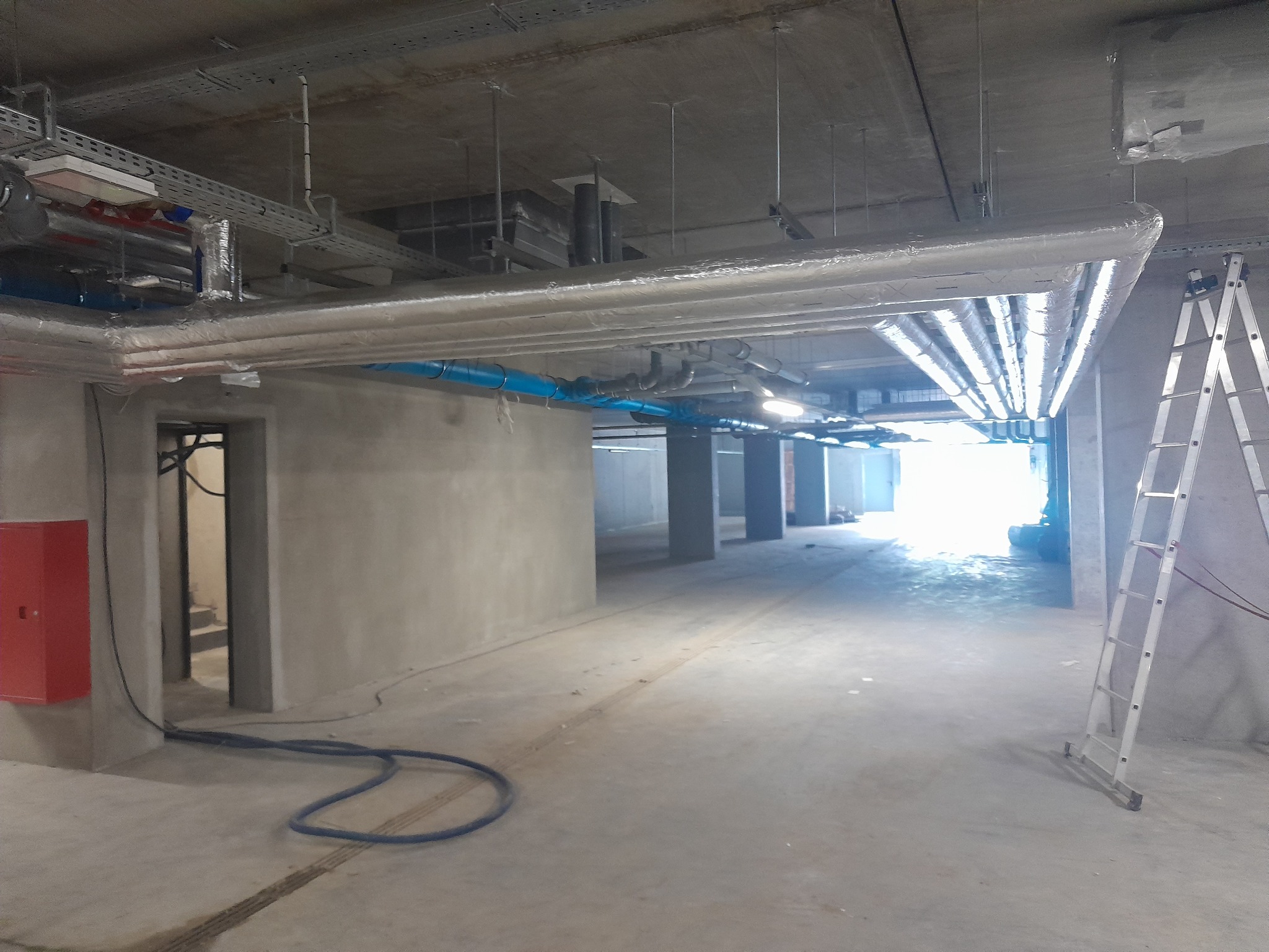 Wnętrze w trakcie instalacji: widoczne rury wentylacyjne pod sufitem, drabina aluminiowa, czerwona szafka hydrantowa i betonowe ściany w surowym stanie.