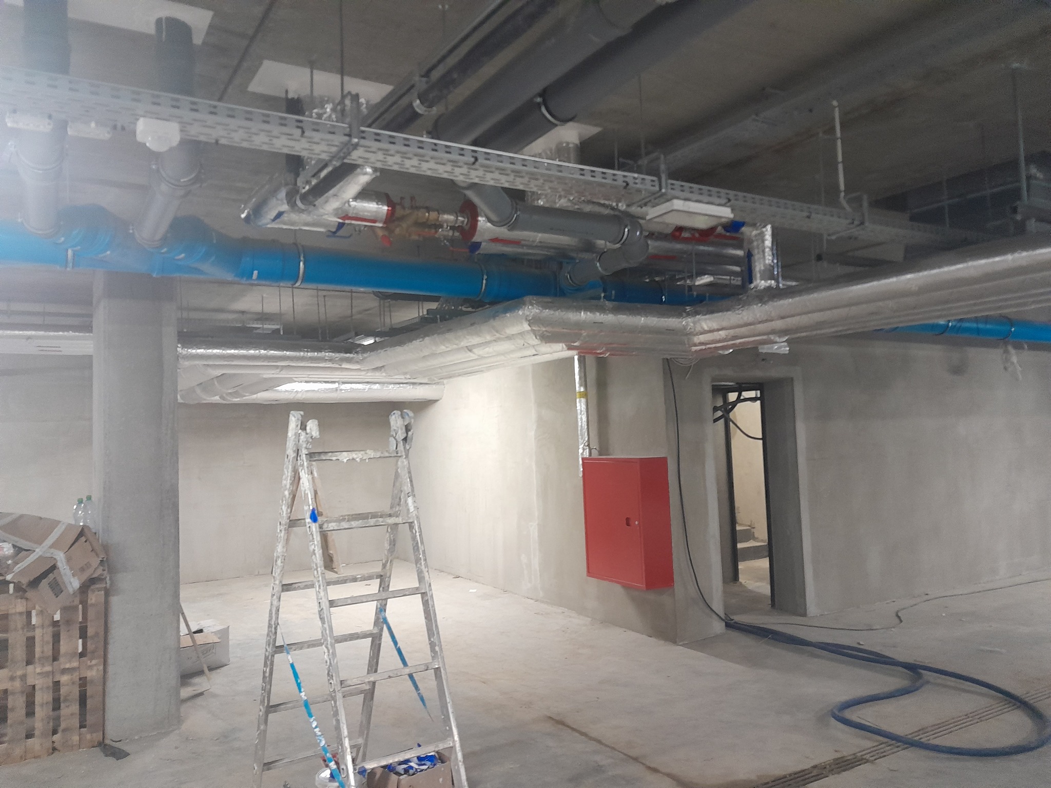 Wnętrze w stanie surowym z widoczną instalacją hydrauliczną i wentylacyjną pod sufitem, drabina malarska i skrzynka hydrantowa.