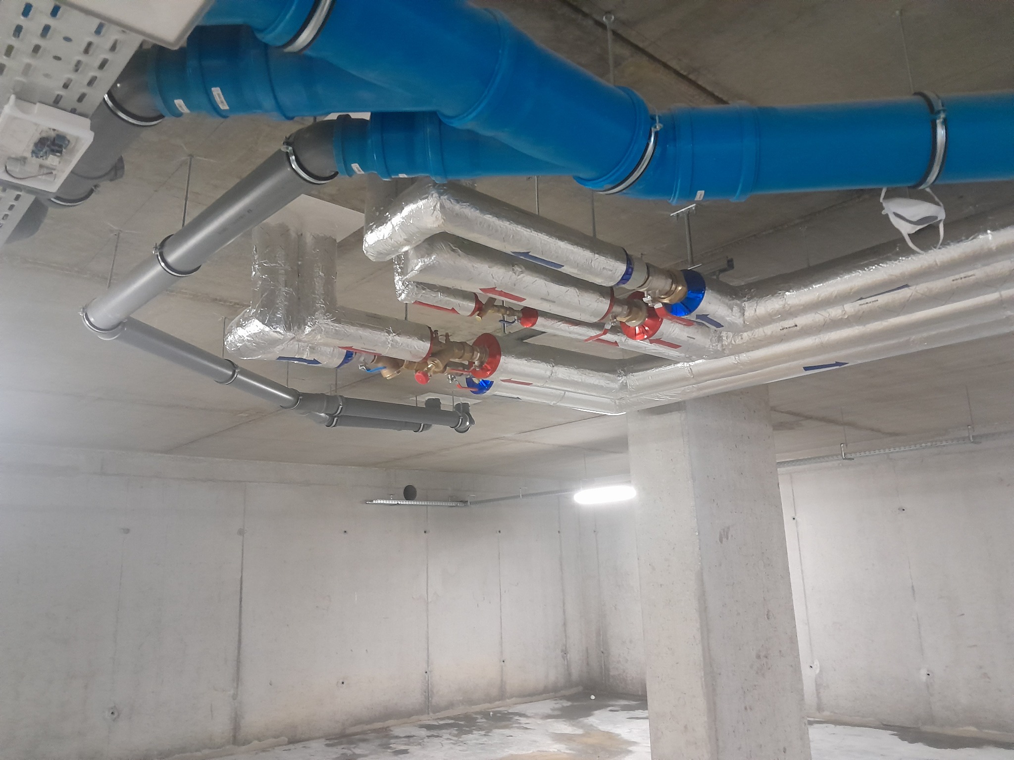 Kompleksowa instalacja rur w piwnicy, rury niebieskie i szare, niektóre izolowane folią, widoczne zawory i mocowania, na jednej z rur wisi maseczka ochronna.