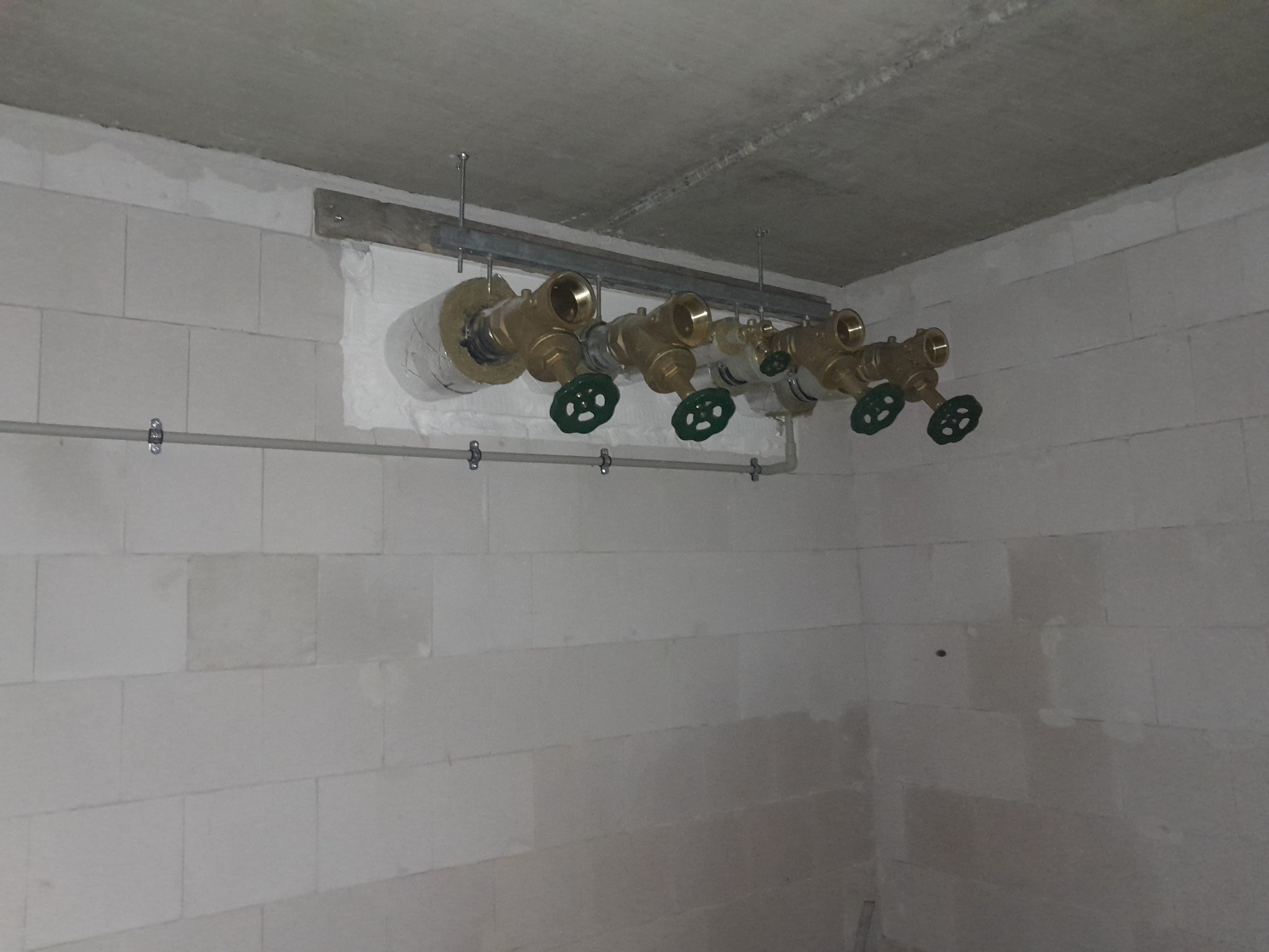 Szereg mosiężnych zaworów z zielonymi pokrętłami, zamontowanych na metalowej szynie pod sufitem w surowym pomieszczeniu z białych bloczków.