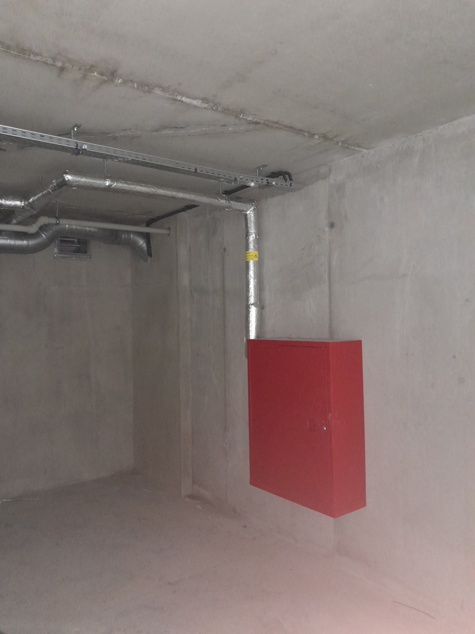 Surowy betonowy pokój z instalacją wentylacyjną pod sufitem i czerwoną szafką na ścianie, widoczne rury owinięte folią aluminiową.