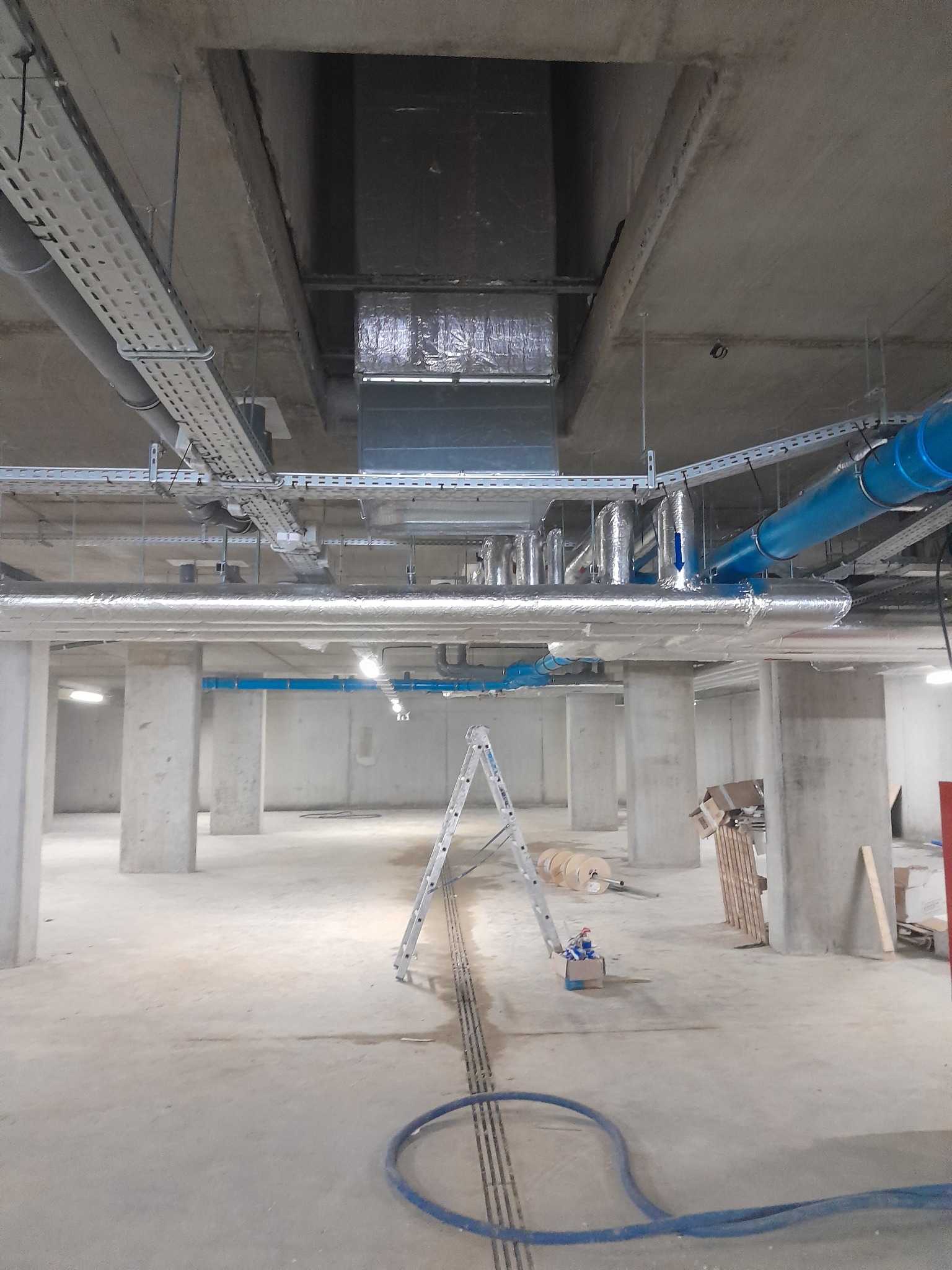 Wnętrze w trakcie instalacji systemów wentylacyjnych i rur, widoczne rury w izolacji, drabina oraz materiały budowlane, surowy betonowy strop i filary.
