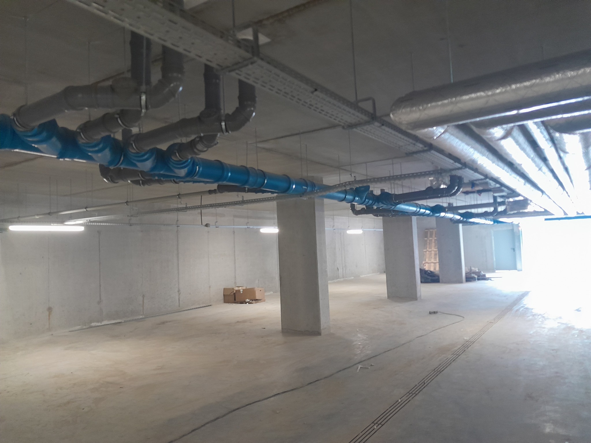 Instalacja rur w garażu podziemnym: niebieskie rury hydrauliczne pod sufitem, szare rury wentylacyjne w izolacji, betonowe filary konstrukcyjne, oświetlenie liniowe LED.