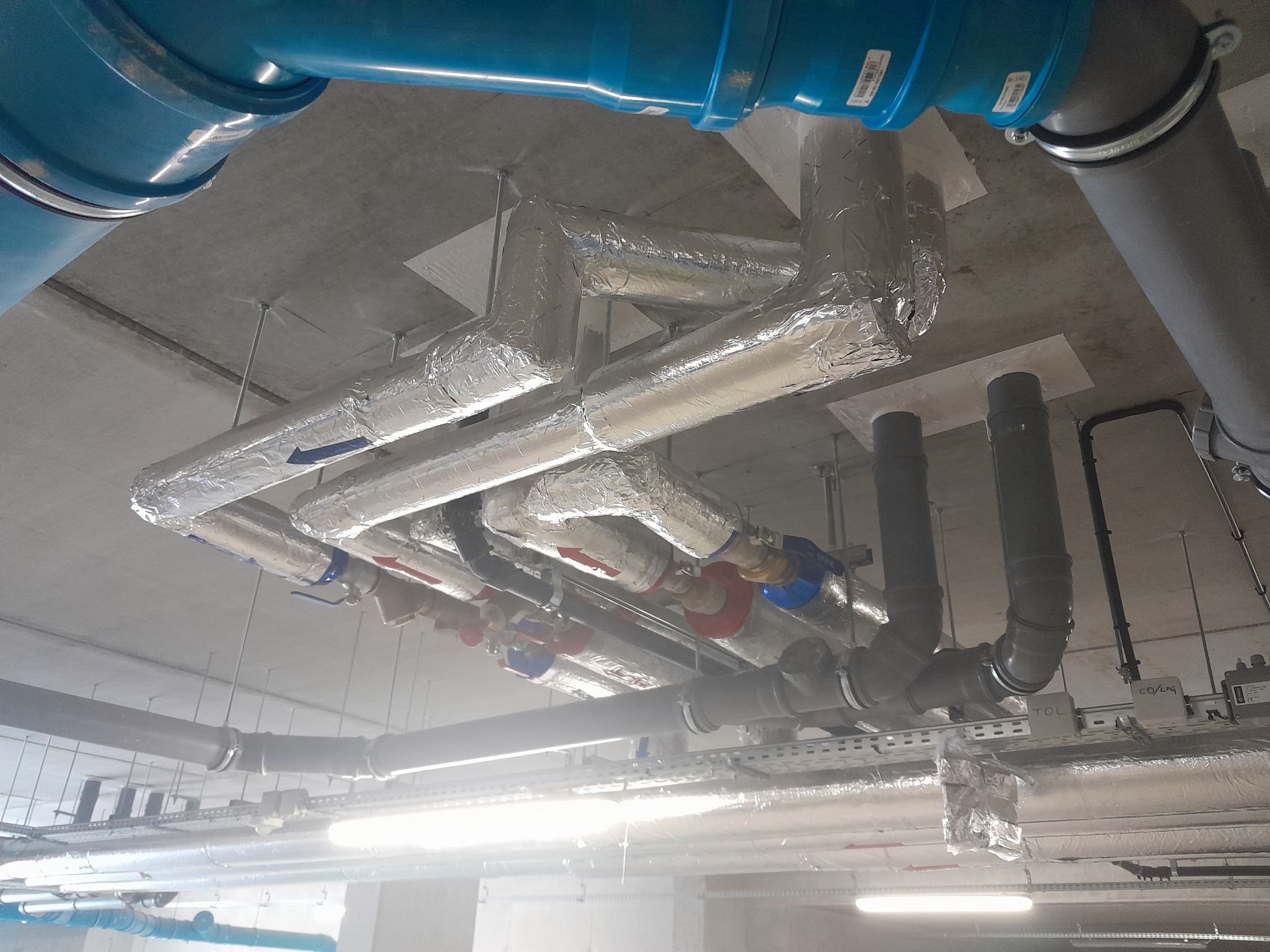 Skomplikowany system rur pod sufitem, niektóre z izolacją termiczną, z widocznymi zaworami i oznaczeniami kierunku przepływu.