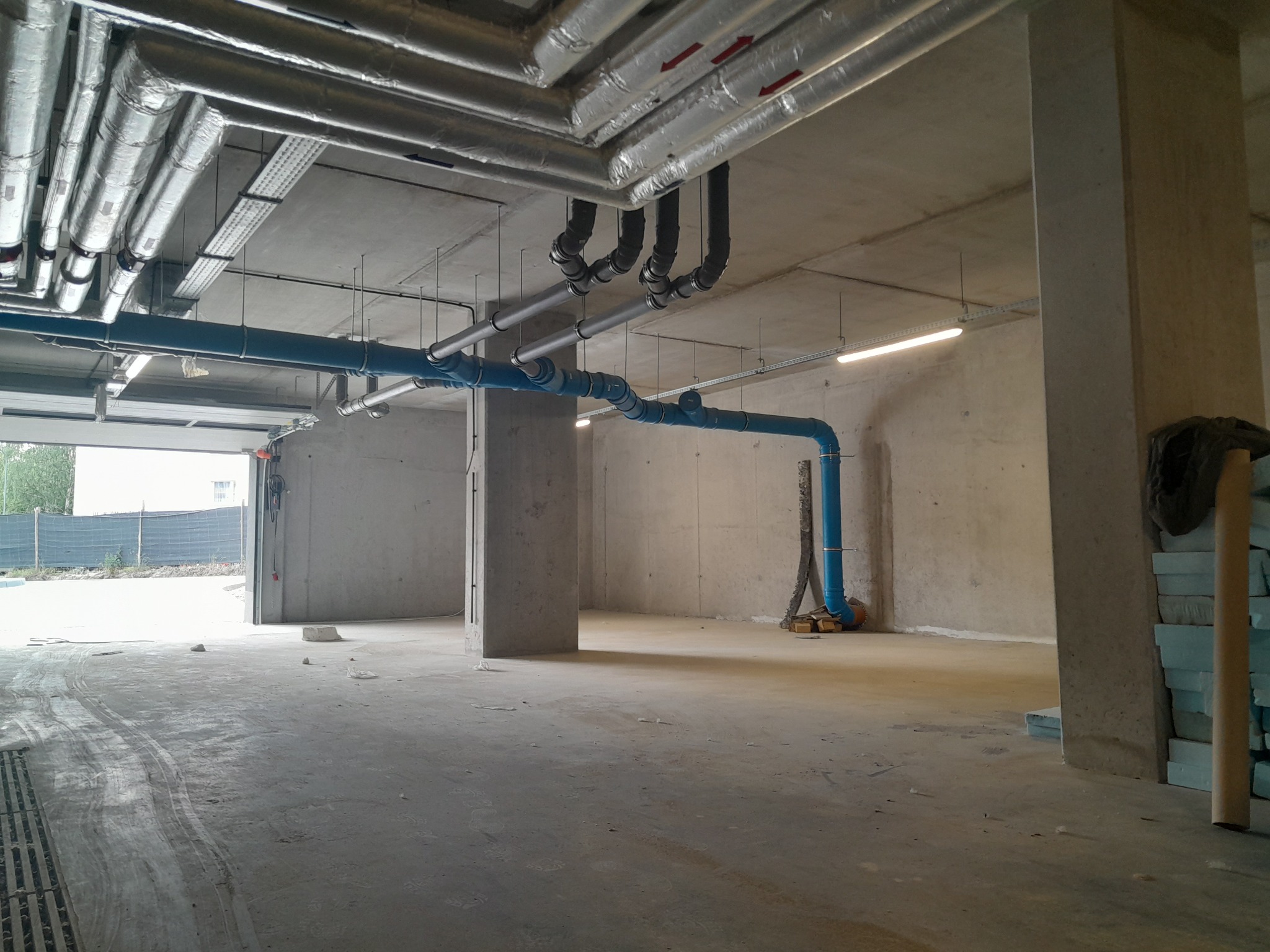 Wnętrze surowego pomieszczenia z widocznymi instalacjami: rury izolowane termicznie pod sufitem, niebieskie rury wodne w pionie i poziomie, betonowe ściany i filary, widoczne ślady po pracach...