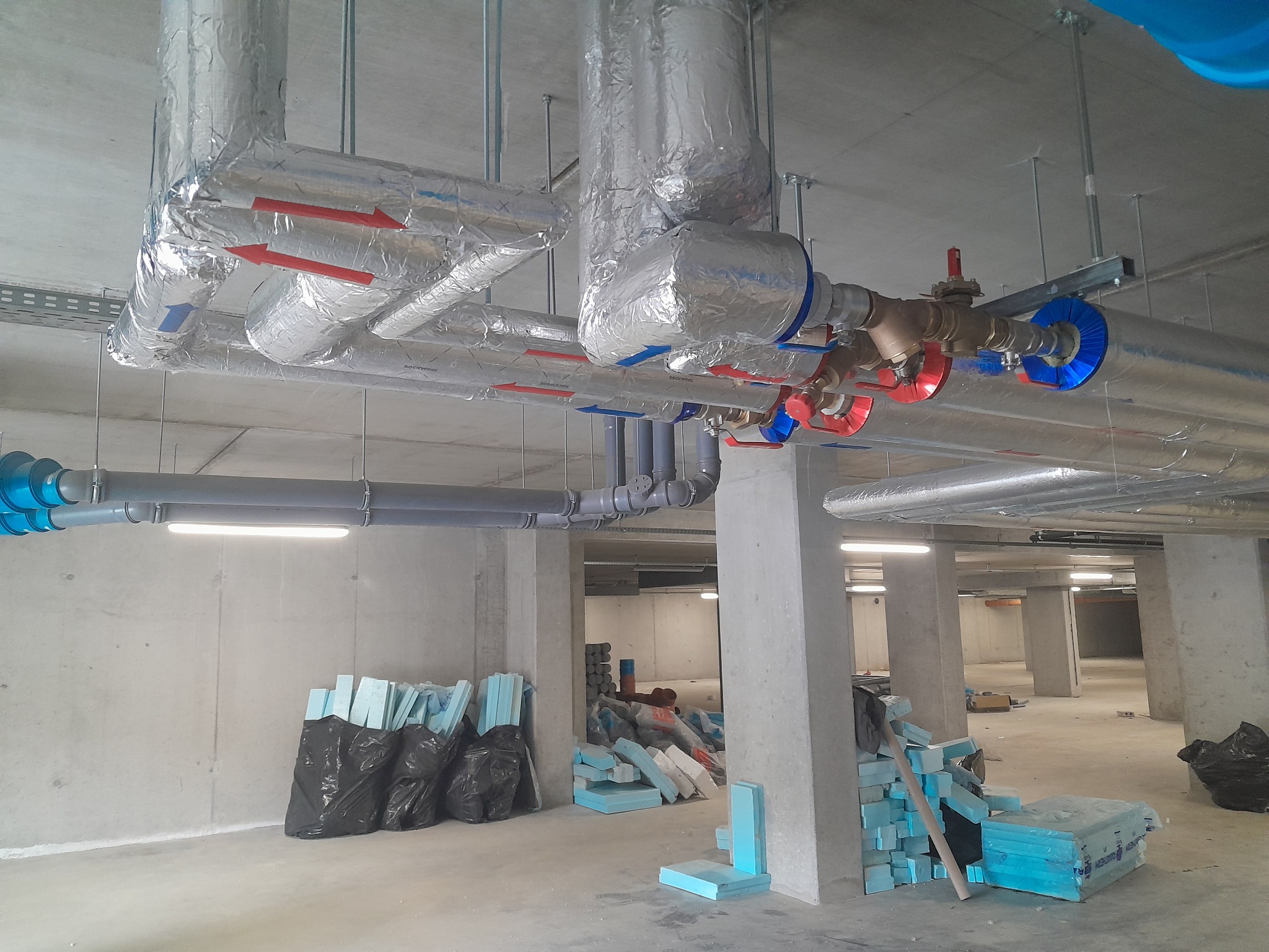 Instalacja grzewcza w garażu podziemnym, rury izolowane folią aluminiową z czerwonymi strzałkami oznaczającymi kierunek przepływu, niebieskie osłony na zaworach, na podłodze materiały izolacyjne...