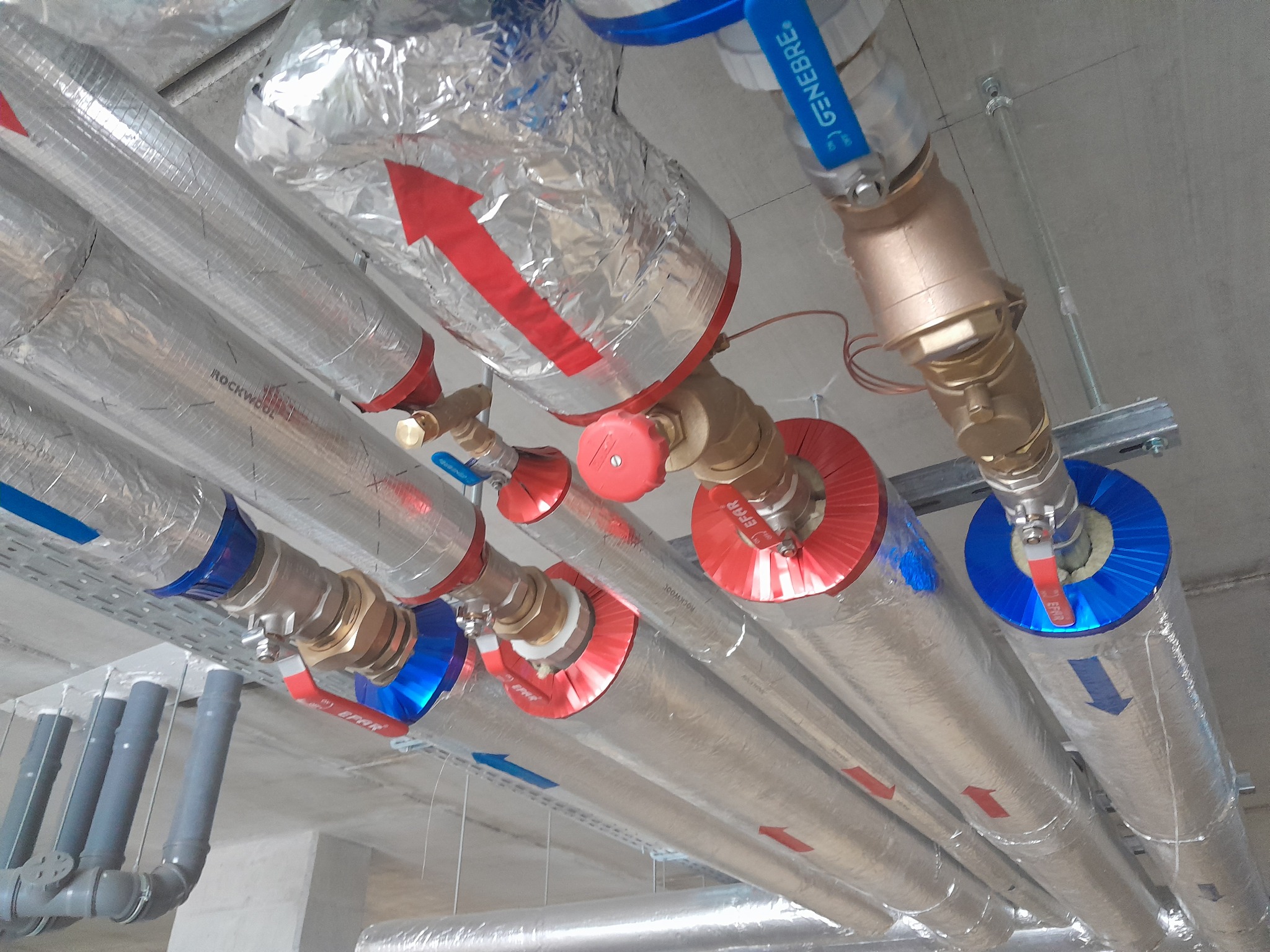 Szereg zaizolowanych rur instalacyjnych pod sufitem, z widocznymi zaworami, strzałkami wskazującymi kierunek przepływu i oznaczeniami Rockwool na izolacji.