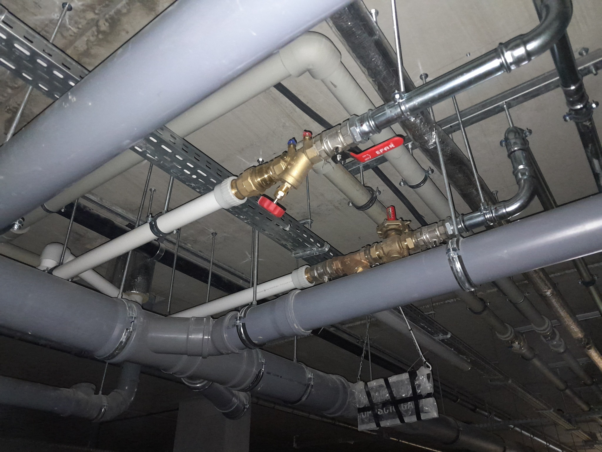 Instalacja hydrauliczna pod sufitem z rur PVC i metalowych, widoczne zawory z czerwonymi pokrętłami, uchwyty mocujące i korytka kablowe.