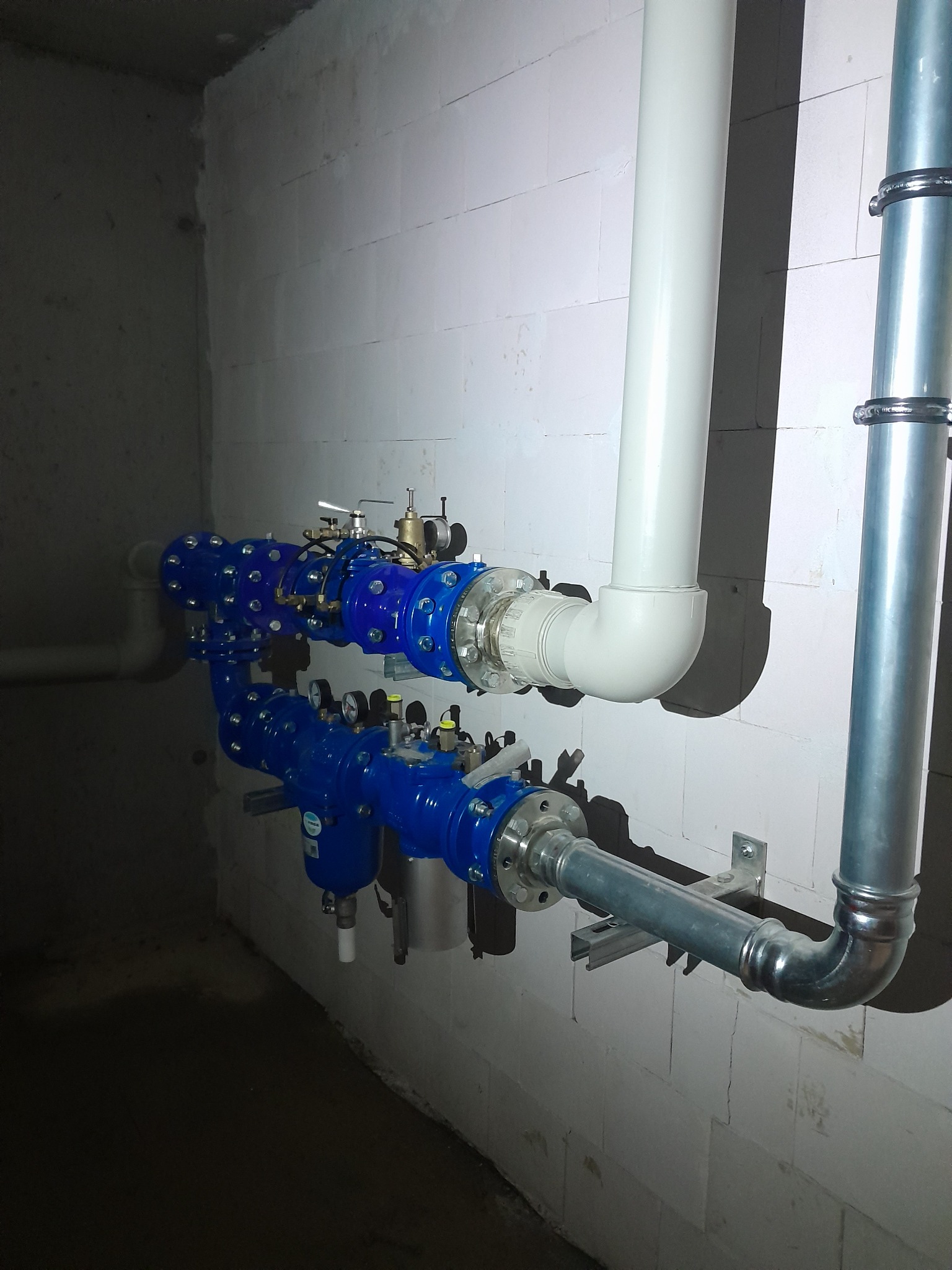 Złożona instalacja rur wodnych w kolorze niebieskim i srebrnym, zamontowana na ścianie z białych bloczków betonowych w piwnicy.