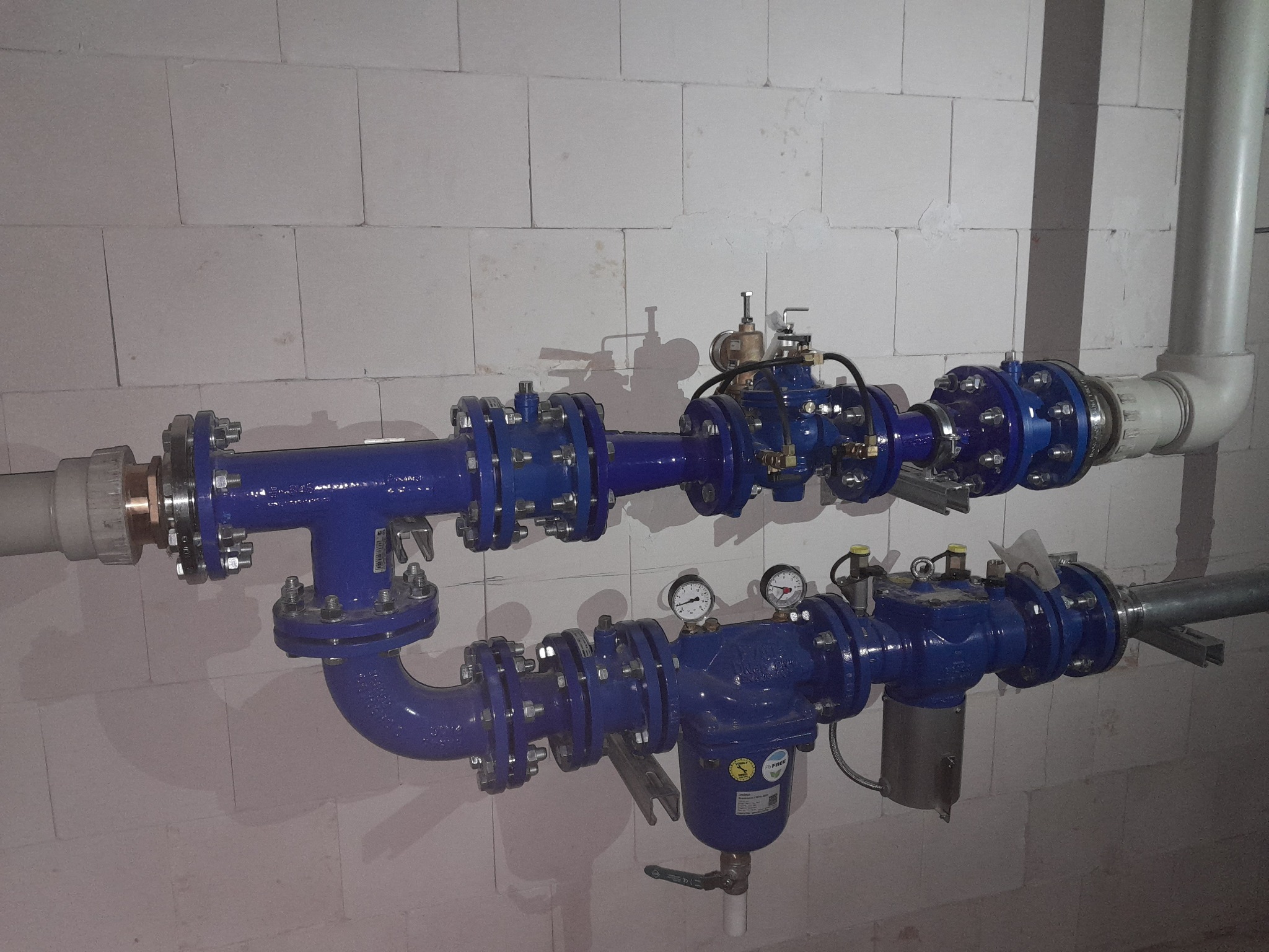 Niebieskie rury hydrauliczne z manometrami i zaworami, mocowane do ściany z białych bloczków betonowych.