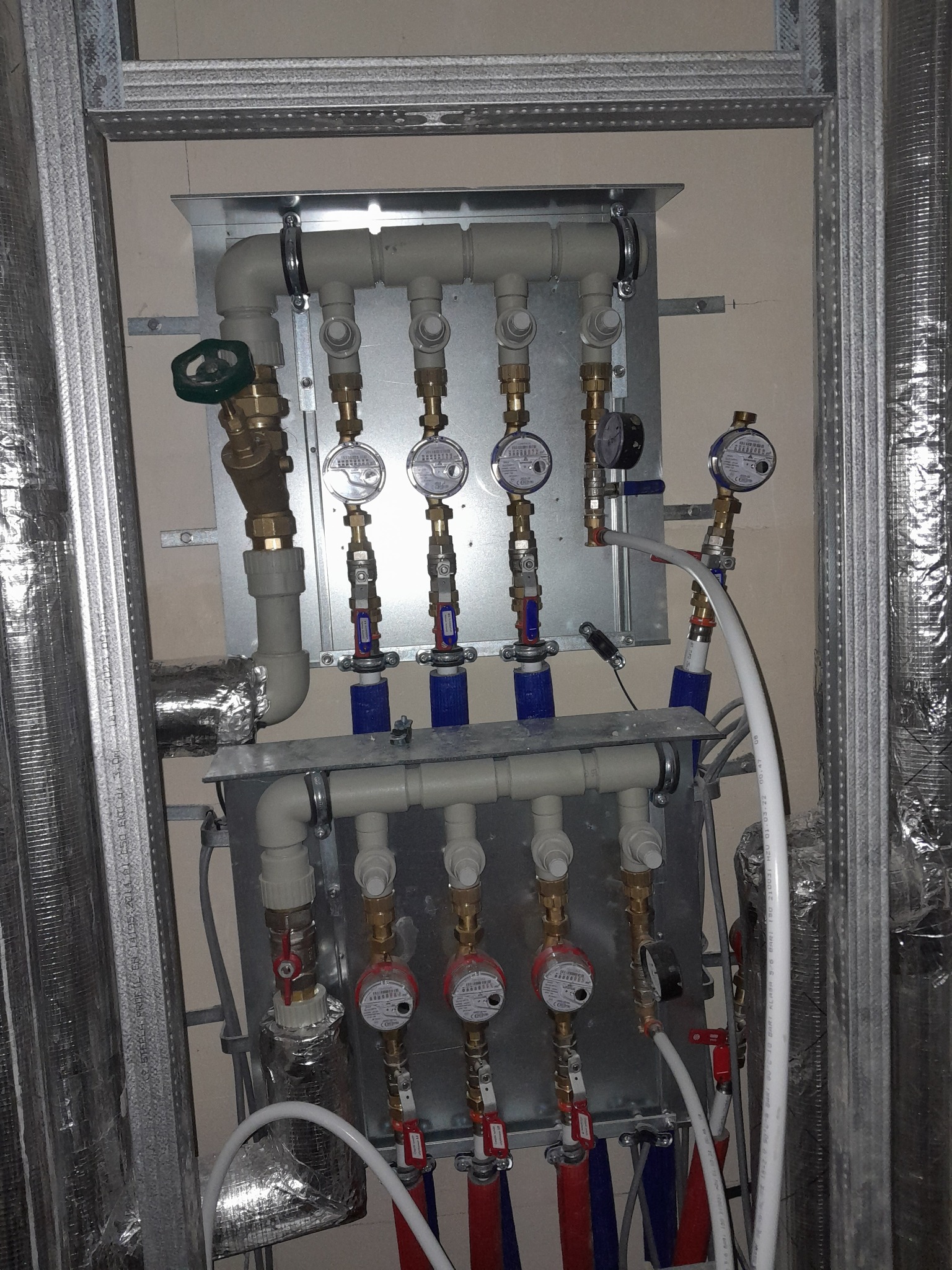Rozdzielacz instalacji wodnej z licznymi zaworami i wodomierzami, zamontowany na metalowej płycie w surowym wnętrzu, widoczne rury i izolacje.