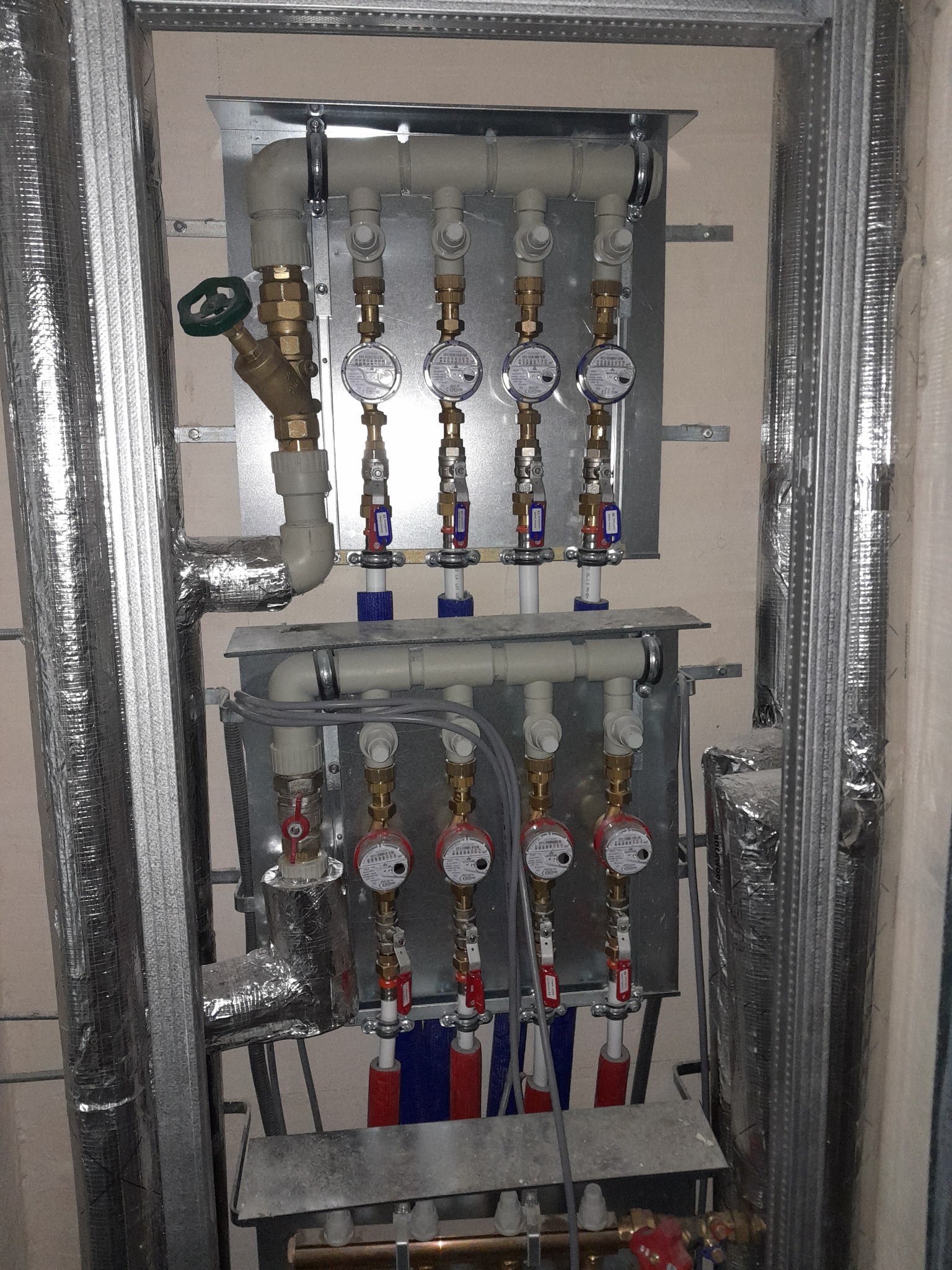Rozdzielacz instalacji grzewczej z zaworami i licznikami ciepła, zamontowany na metalowej płycie, widoczne rury izolowane folią aluminiową oraz fragment konstrukcji nośnej.