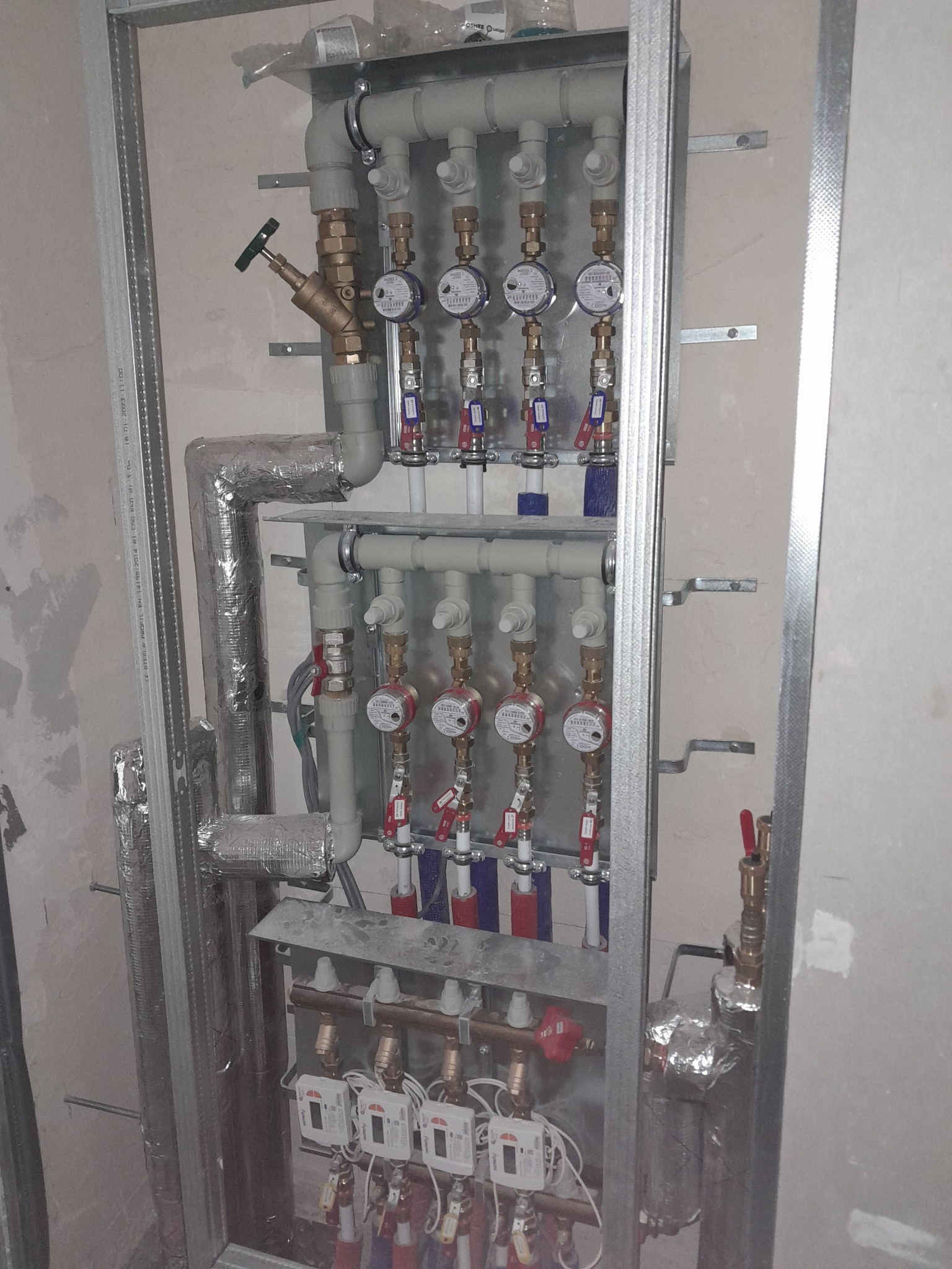 Rozdzielacz instalacji grzewczej z licznikami ciepła, rury izolowane folią aluminiową, zamontowany w metalowej konstrukcji szkieletowej na tle surowej ściany.