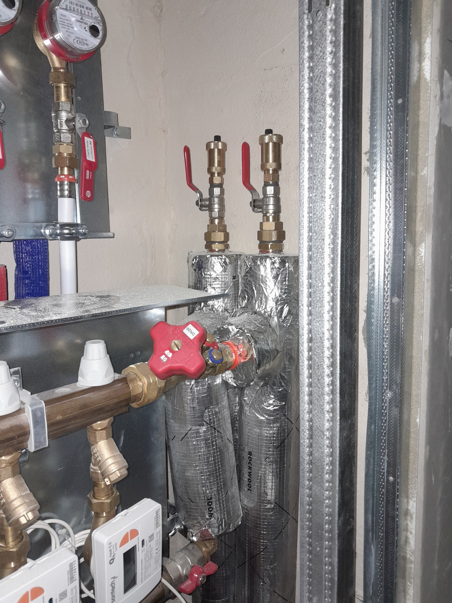 Węzeł cieplny w kotłowni: izolowane rury grzewcze z zaworami, liczniki ciepła Sanitech, wodomierz Apator, instalacja na metalowym stelażu przy ścianie.