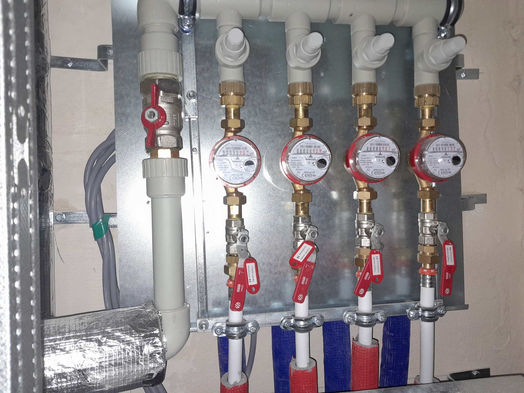 Rozdzielacz instalacji grzewczej z czterema licznikami ciepła, zaworami i izolacją termiczną rur, zamontowany na metalowej płycie.