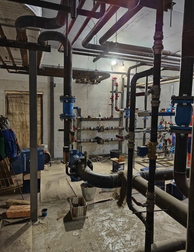 Piwnica z widocznymi rurami instalacji hydraulicznej i grzewczej, z niebieskimi zaworami i pompą Grundfos, w surowym stanie, z widocznymi śladami prac instalacyjnych.