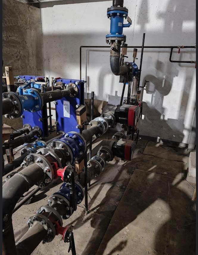 Węzeł cieplny: widok na skomplikowany system rur, zaworów i pomp w kotłowni, z niebieskim wymiennikiem ciepła płytowym po lewej stronie i dwoma pompami obiegowymi z czerwonymi elementami, całość...