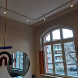 Wnętrze pokoju z zamontowanym oświetleniem szynowym na suficie, okrągłym lustrem na ścianie i dużym oknem łukowym z widokiem na budynek mieszkalny.