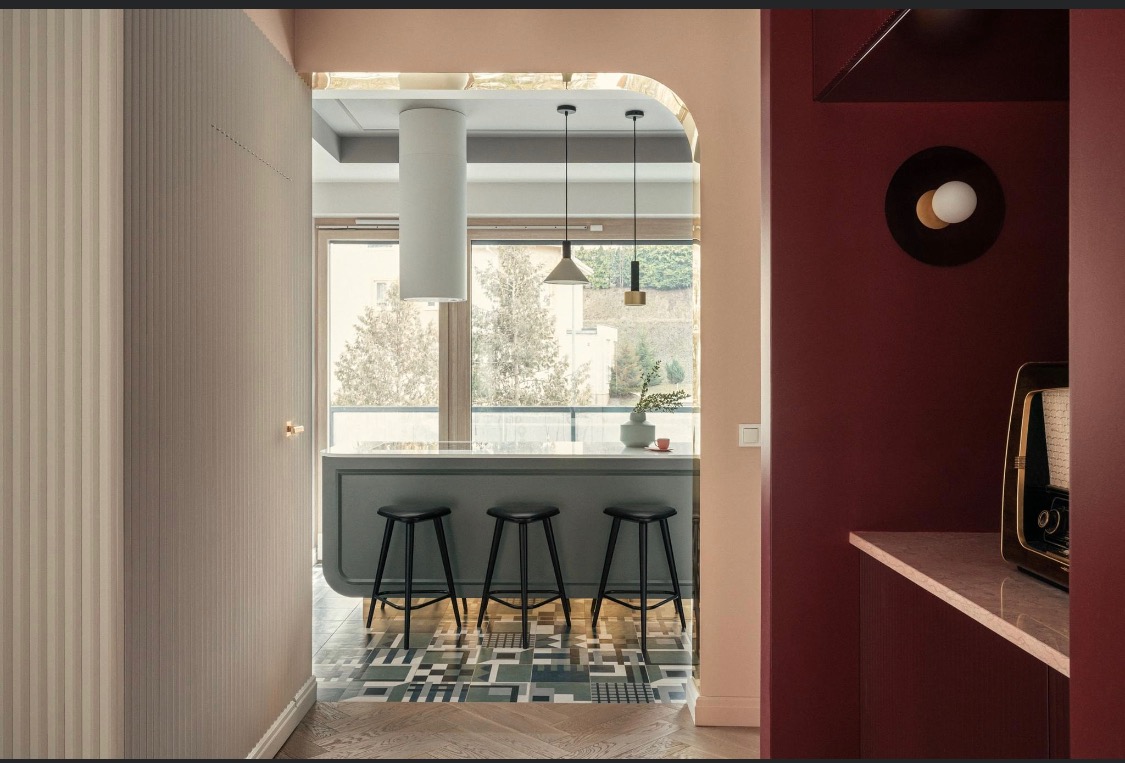 Wnętrze mieszkania z widokiem na kuchnię z trzema czarnymi stołkami barowymi i wzorzystą podłogą, widoczne elementy wykończeniowe ścian w różnych kolorach i fakturach, oraz stara radioodbiornik...