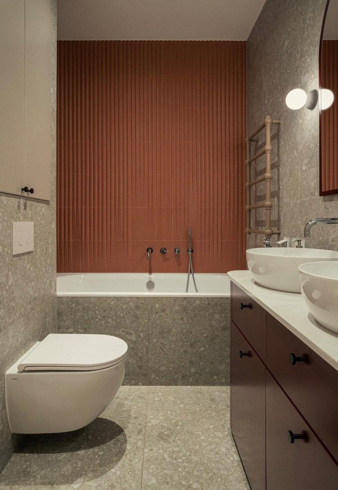 Nowoczesna łazienka z wanną obudowaną płytkami lastryko, umywalkami nablatowymi i ścianą z pionowych, brązowych płytek dekoracyjnych.