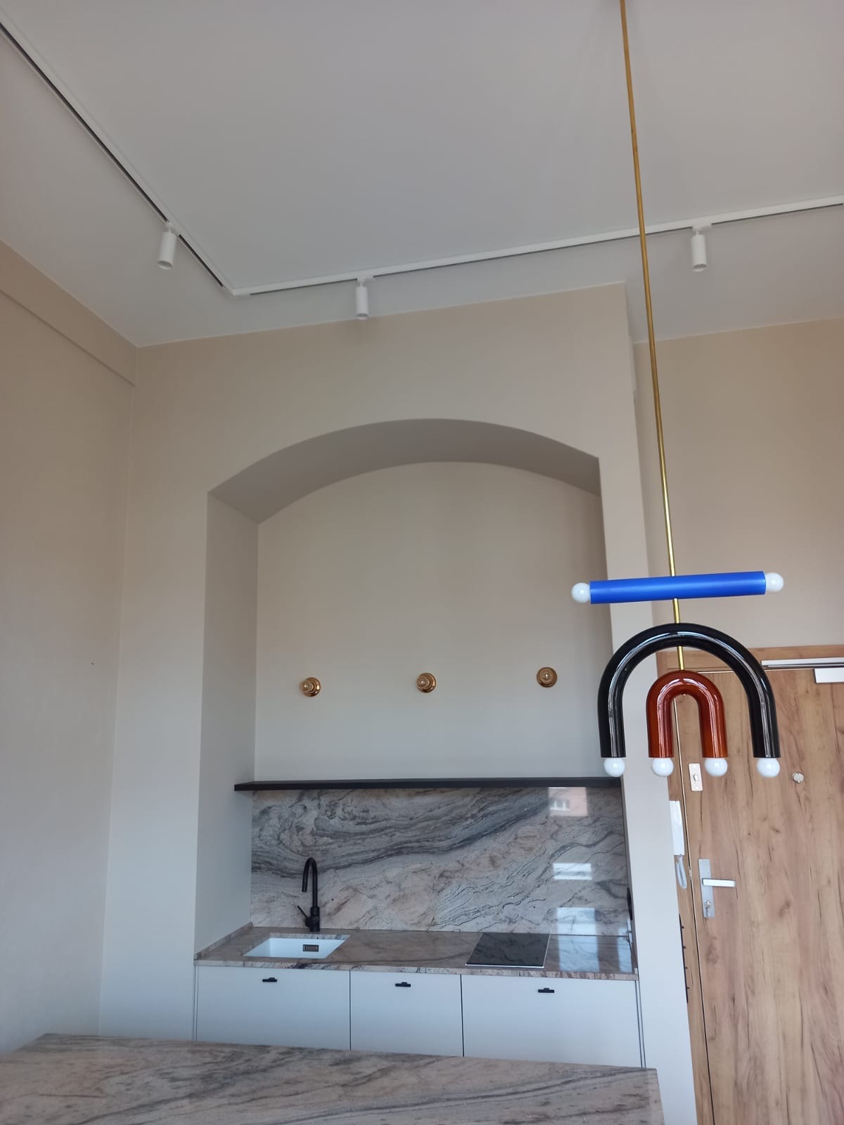 Nowoczesna kuchnia z białymi szafkami i blatem z marmuru, czarną armaturą, oświetleniem szynowym i designerską lampą wiszącą w kształcie łuków.