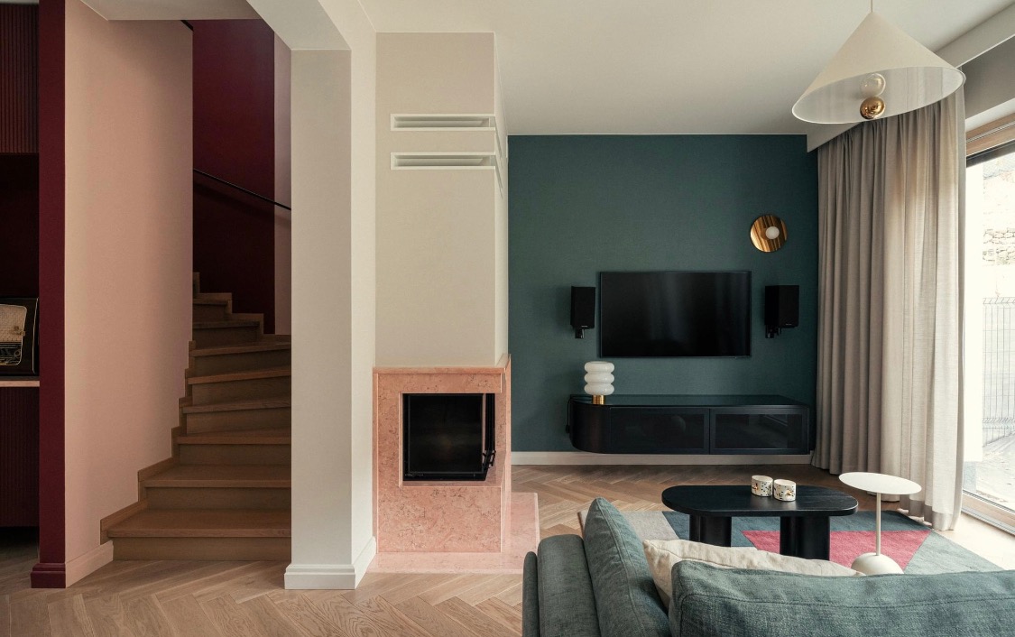 Nowoczesny salon z kominkiem obłożonym różowym marmurem, schodami z jasnego drewna i ścianami w odcieniach zieleni i różu, oświetlony wiszącą lampą z białym kloszem.