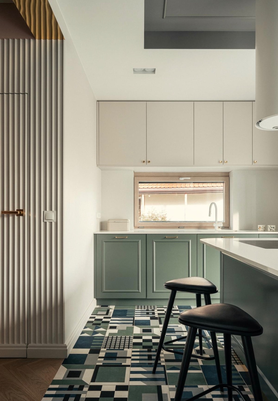 Nowoczesna kuchnia z zielonymi szafkami, geometryczną podłogą i dwoma czarnymi stołkami barowymi.