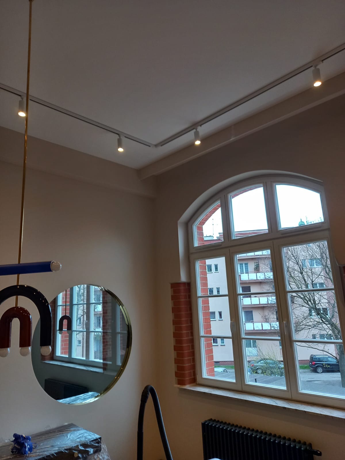 Wnętrze pokoju z zamontowanym oświetleniem szynowym na suficie, okrągłym lustrem na ścianie i dużym oknem łukowym z widokiem na budynek mieszkalny.