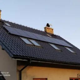 Panele słoneczne zamontowane na ciemnym, spadzistym dachu domu jednorodzinnego z oknami dachowymi i kominem z cegły, sfotografowane w pogodny dzień.