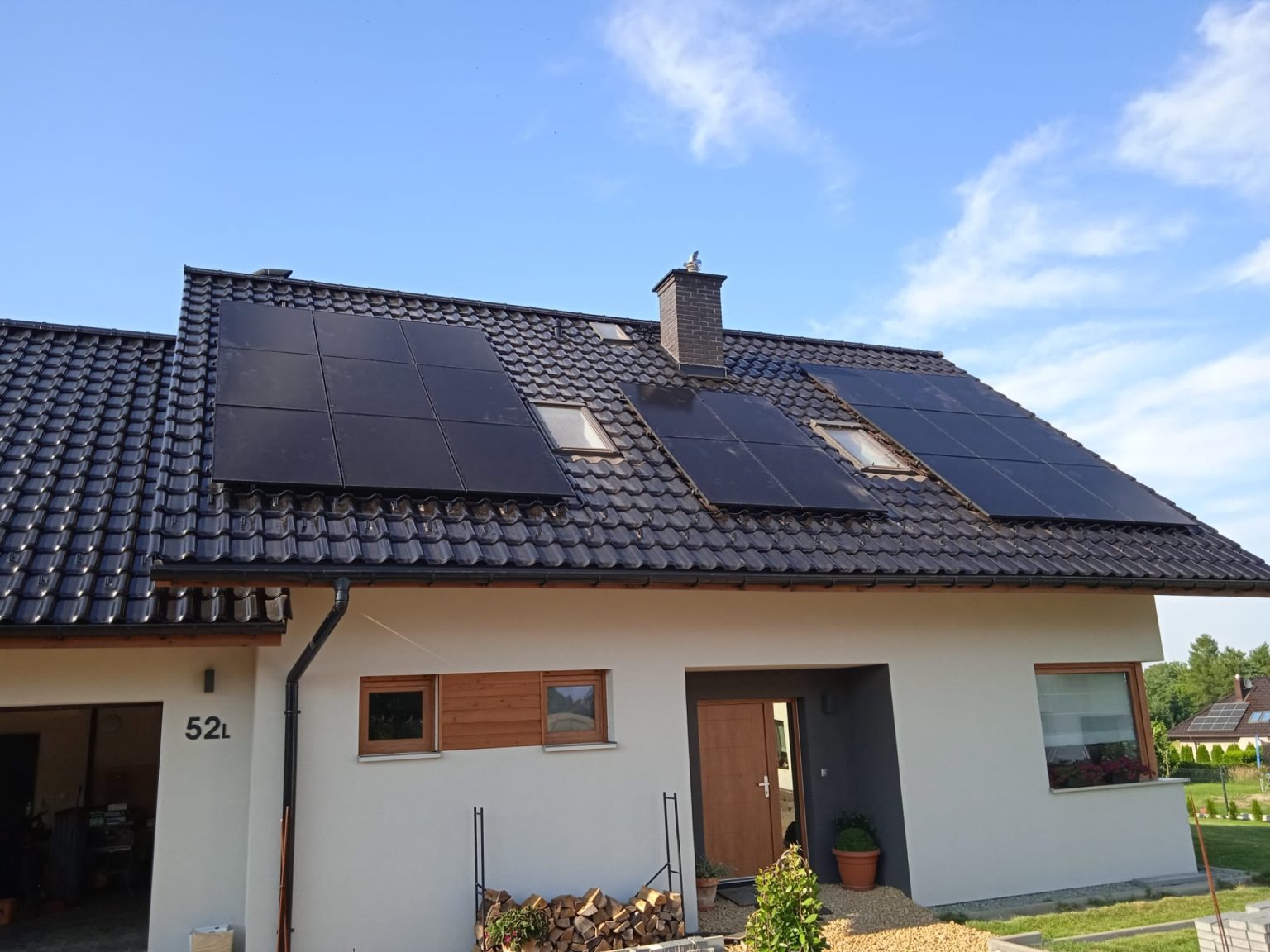 Czarny dach pokryty panelami fotowoltaicznymi na nowoczesnym domu jednorodzinnym z jasną elewacją i oknami dachowymi, widok z dołu w słoneczny dzień.