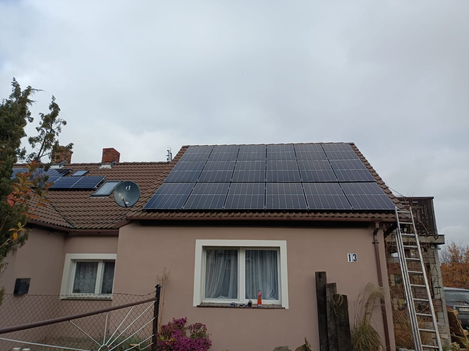 Panele słoneczne zamontowane na dachu domu jednorodzinnego z numerem 13, widoczna drabina oparta o budynek, pochmurne niebo w tle.